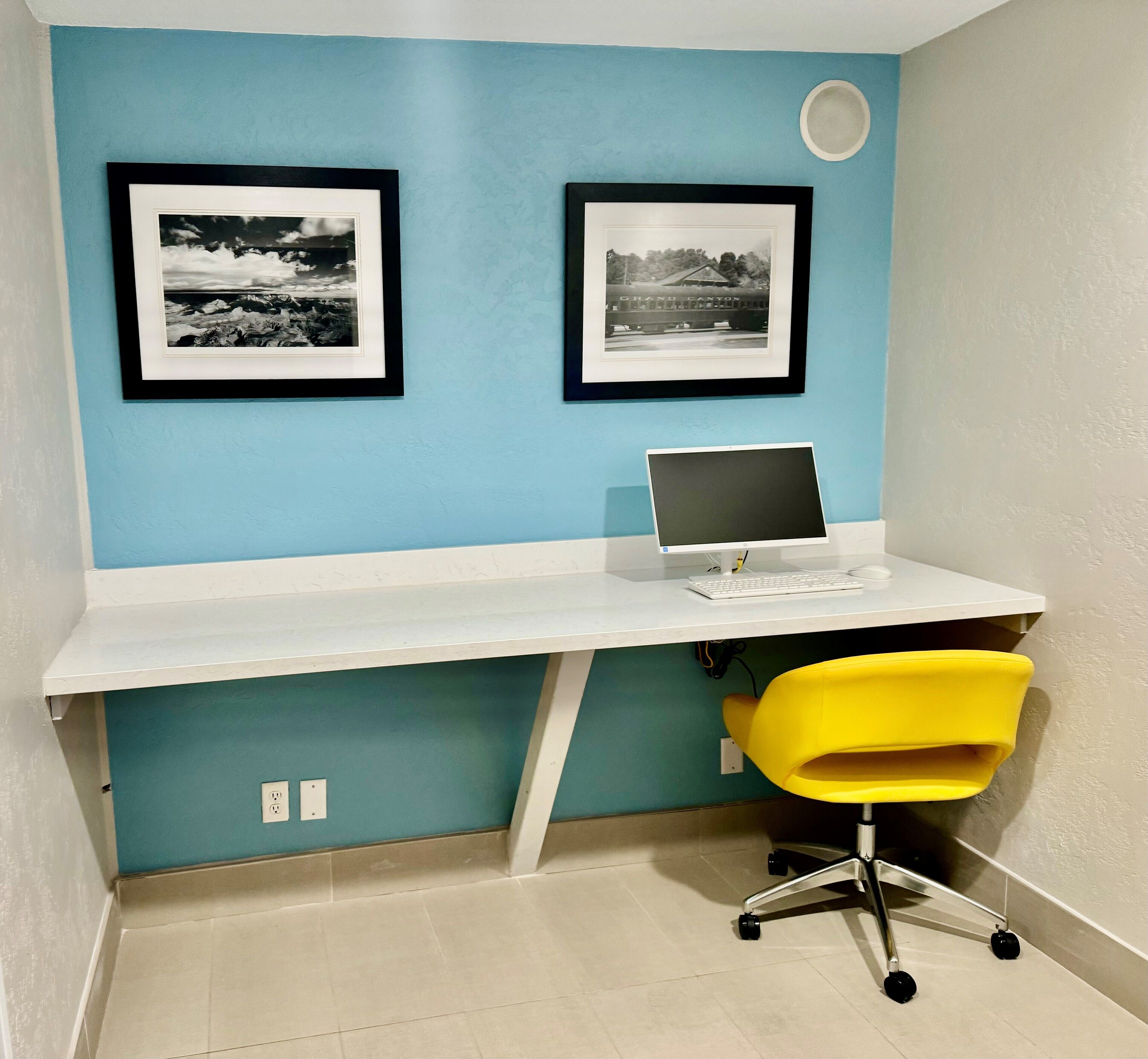 A small office space with a white desk, a laptop, a yellow chair, and two framed black and white photographs on the wall.