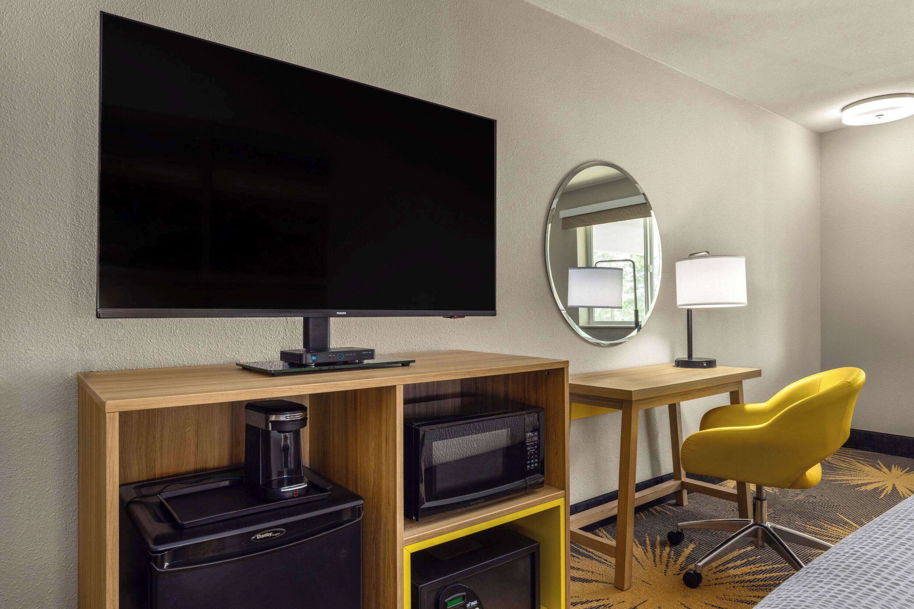 Ein Hotelzimmer mit Flachbildfernseher, einer Kaffeemaschine, einer Mikrowelle, einem Schreibtisch, einem Stuhl und einem Bett.