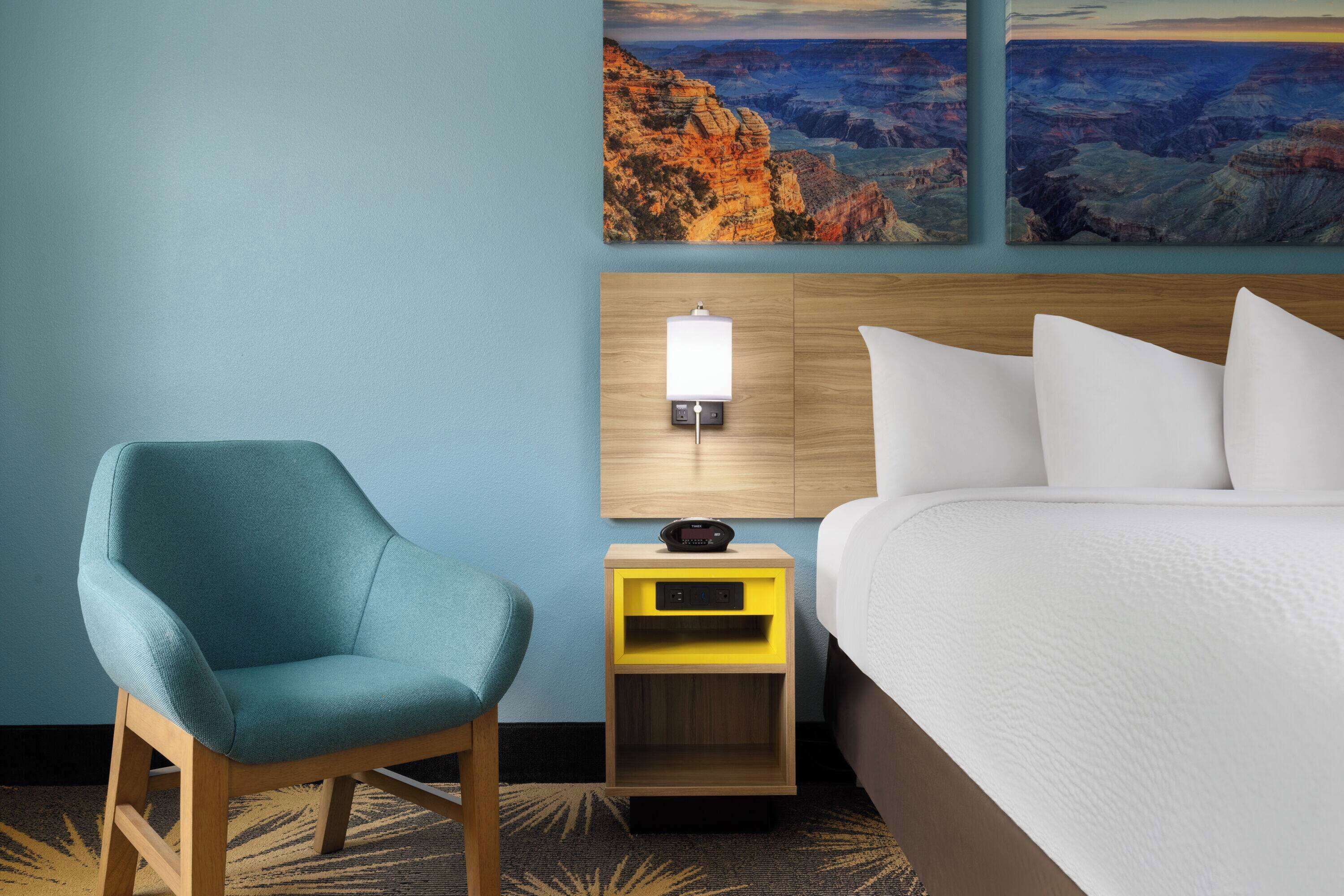 A hotel room with a bed, a chair, a nightstand with a lamp, and two paintings of a canyon.