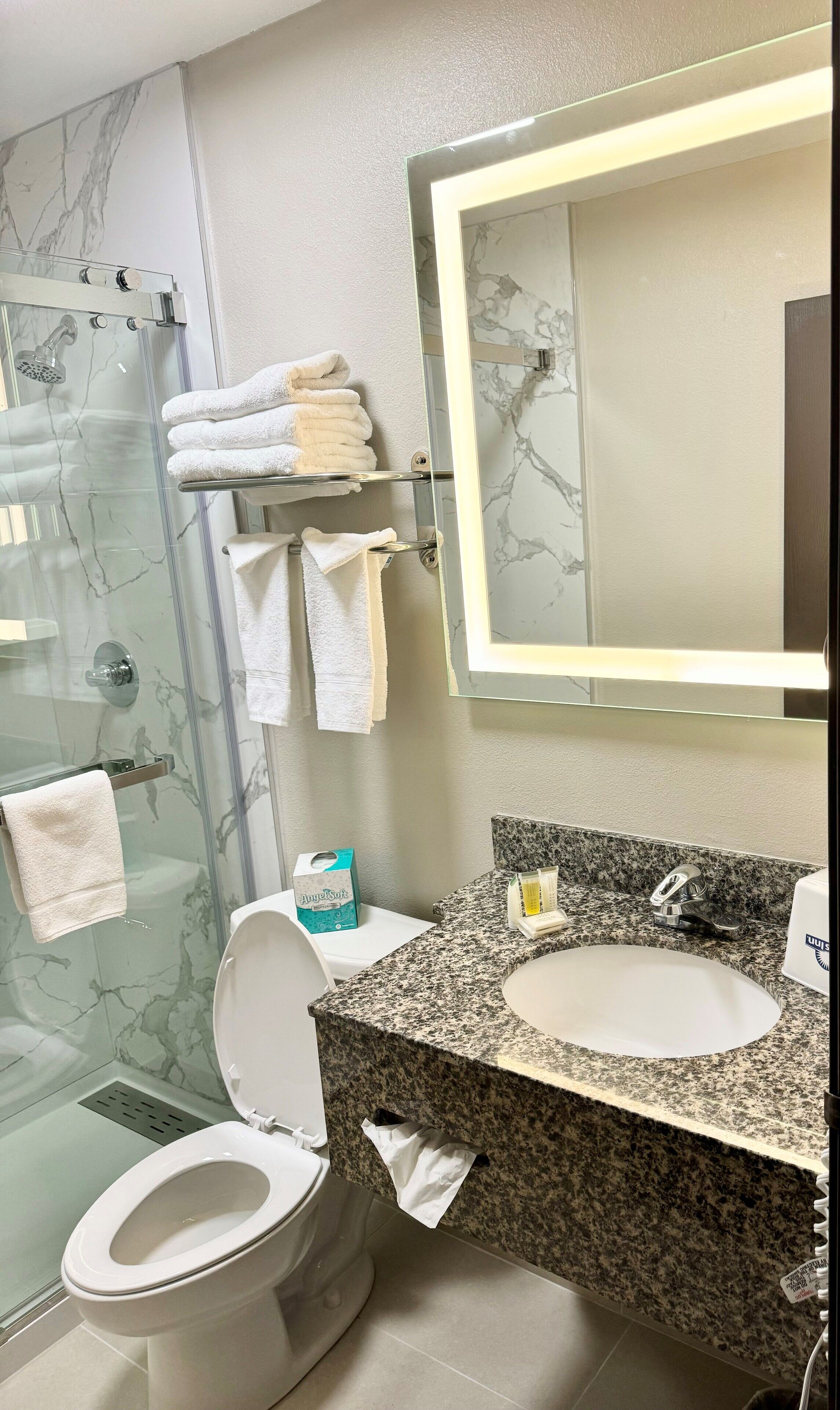 Un baño con ducha revestida de mármol, inodoro, lavamanos con encimera de granito y espejo con luz.