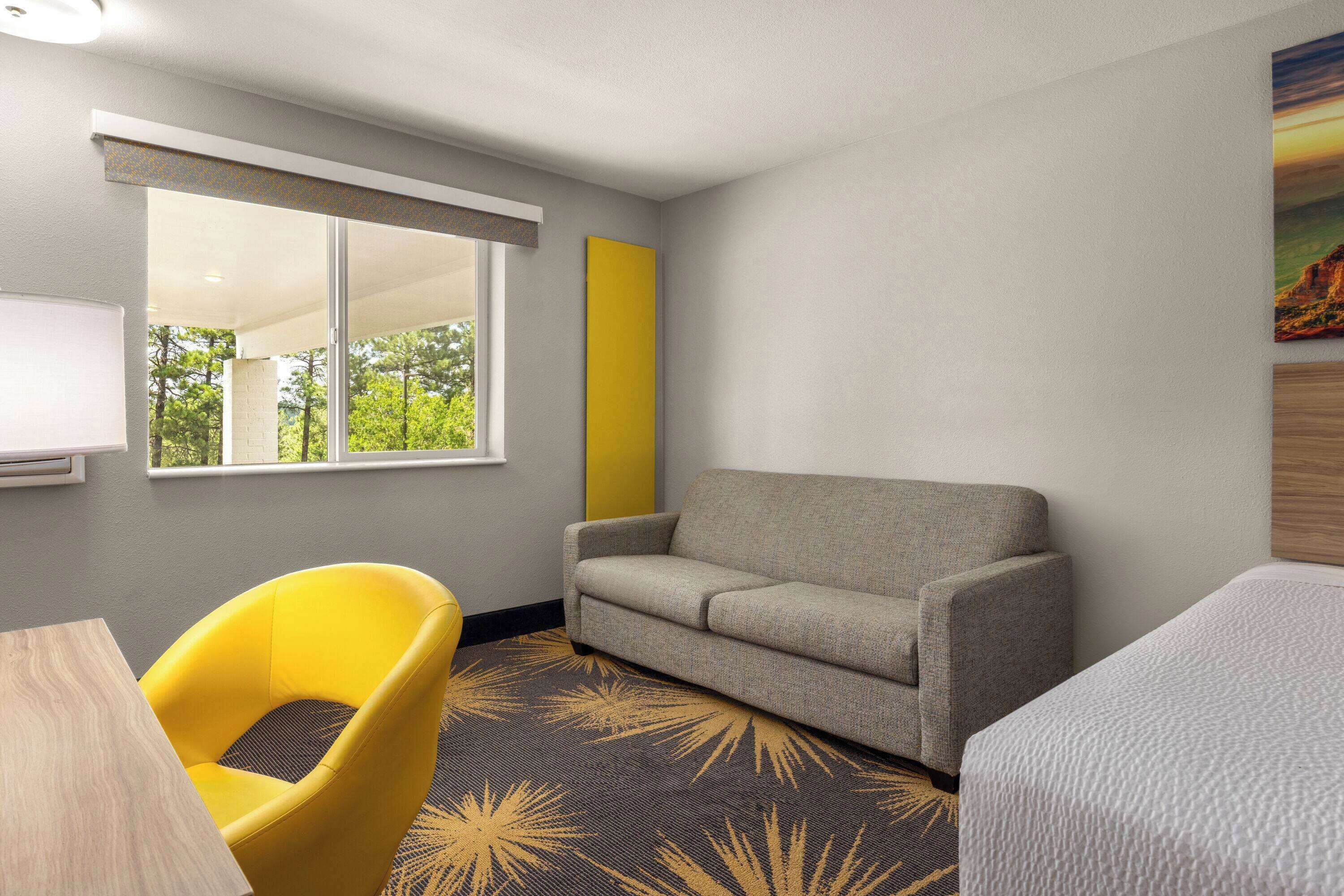 Une chambre d’hôtel avec un canapé, un fauteuil jaune, un lit, une fenêtre donnant sur des arbres et un tableau au mur.