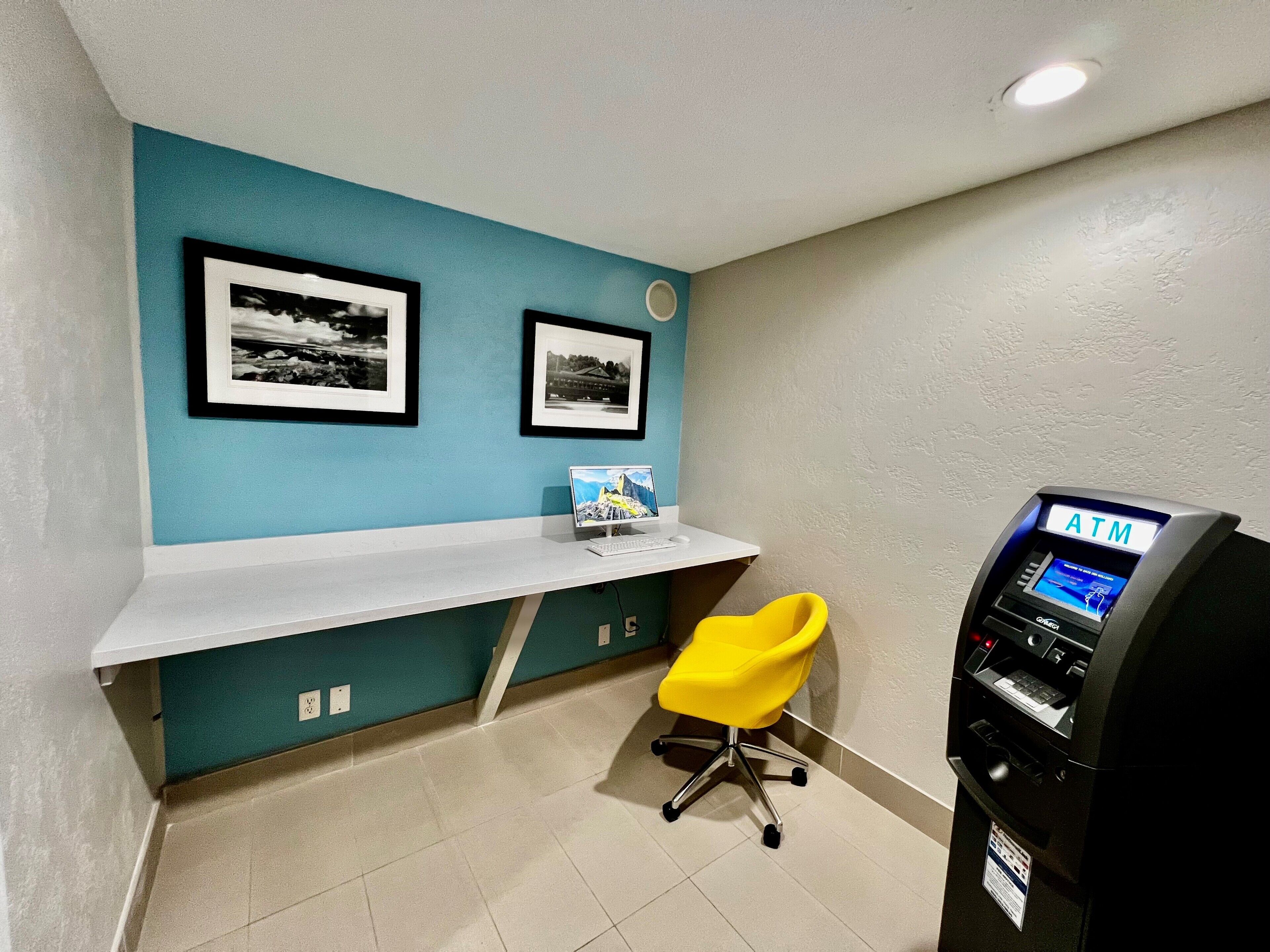 Een kleine kantoorruimte met een bureau, twee ingelijste foto's, een gele stoel en een geldautomaat.