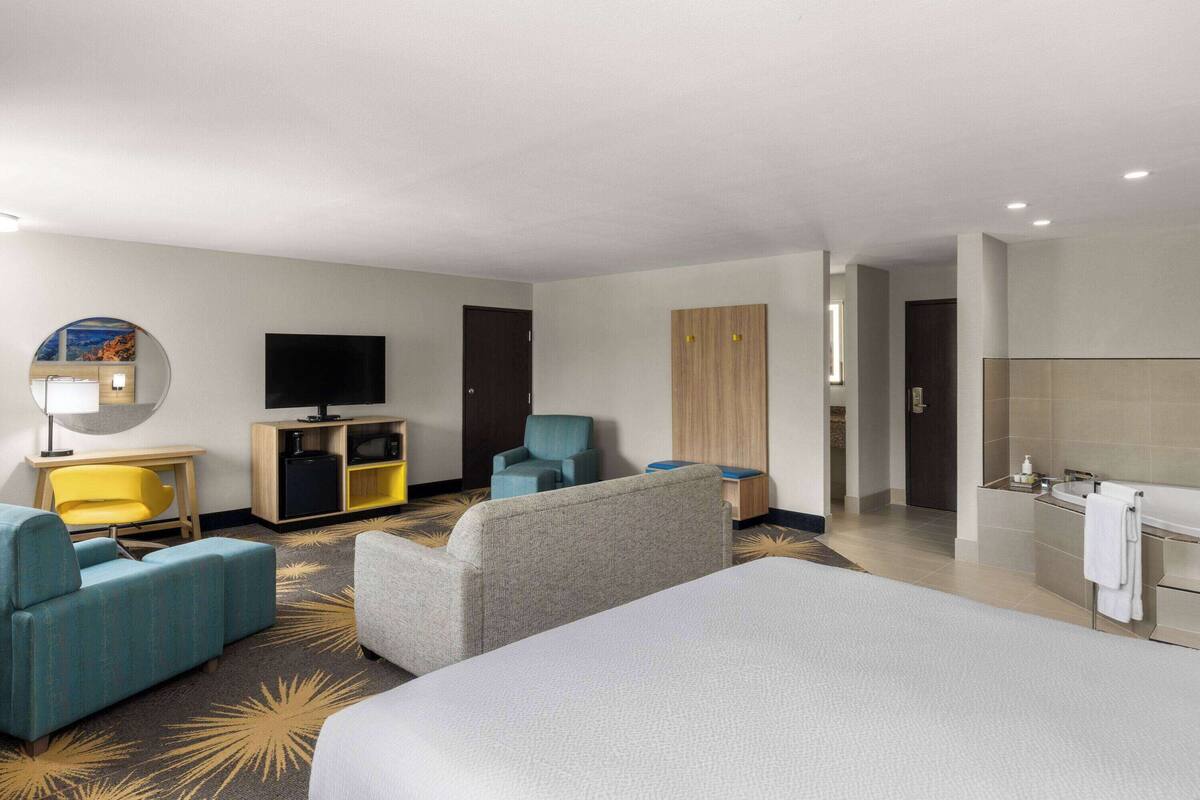 A hotel room with a large bed, a sofa, a TV, a desk, and a bathroom with a sink and mirror.
