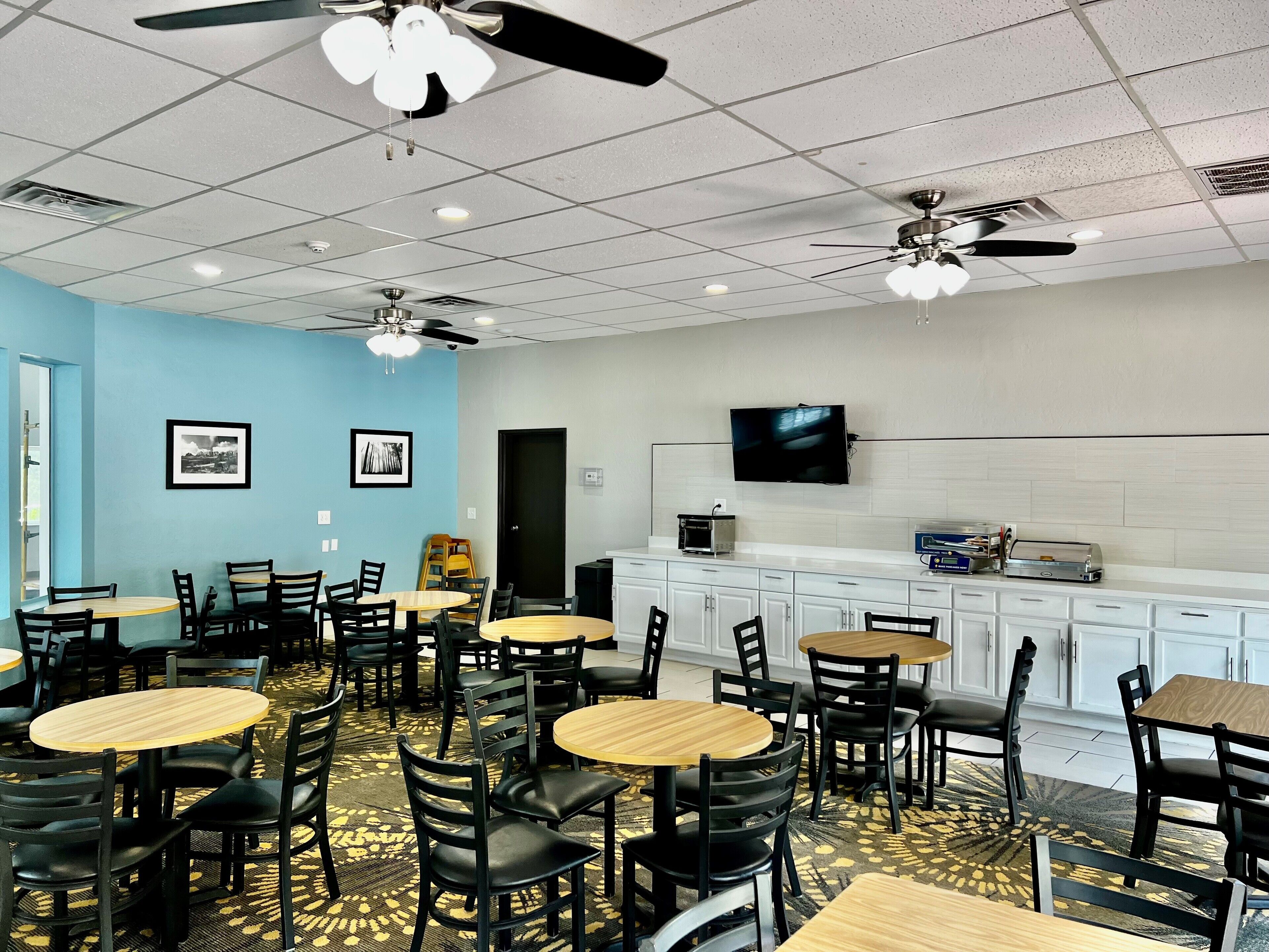 A conference room with multiple tables and chairs, ceiling fans, a flat-screen TV, and a kitchenette area.