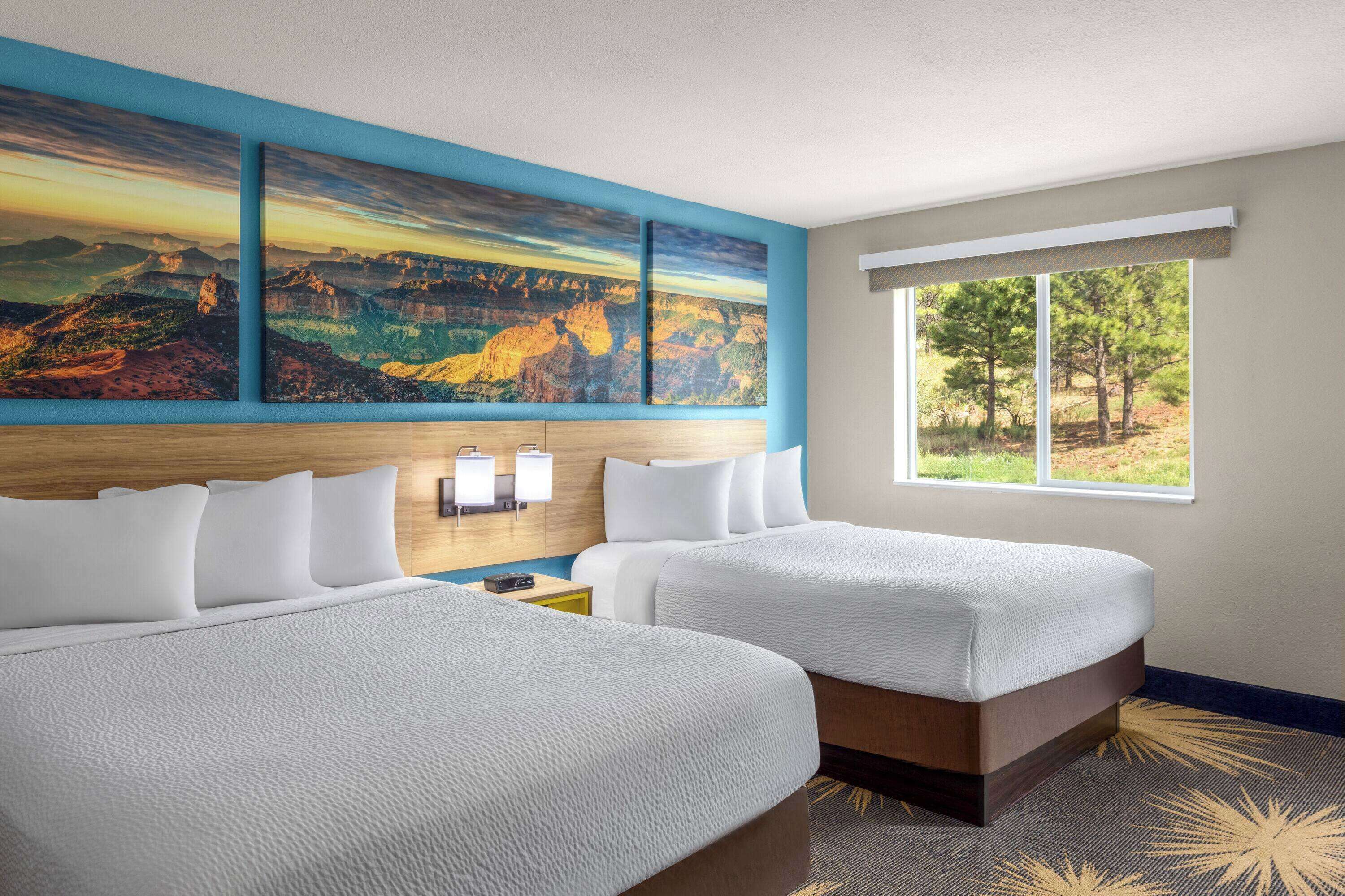 A hotel room with two beds, a large painting of a canyon, a window with a view of trees, and a nightstand with a lamp.