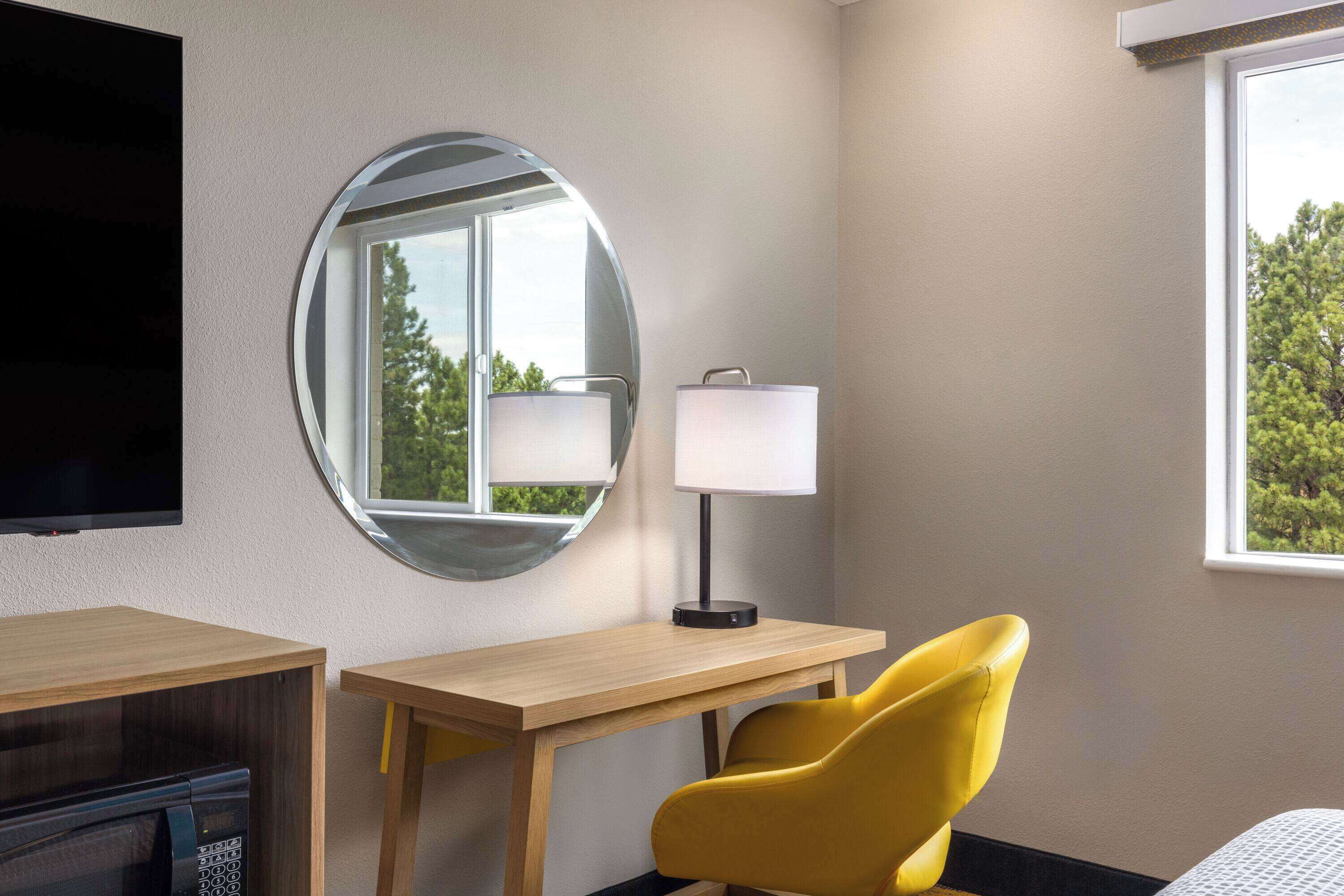 A hotel room with a wooden desk, a yellow chair, a round mirror, a lamp, and a TV mounted on the wall.