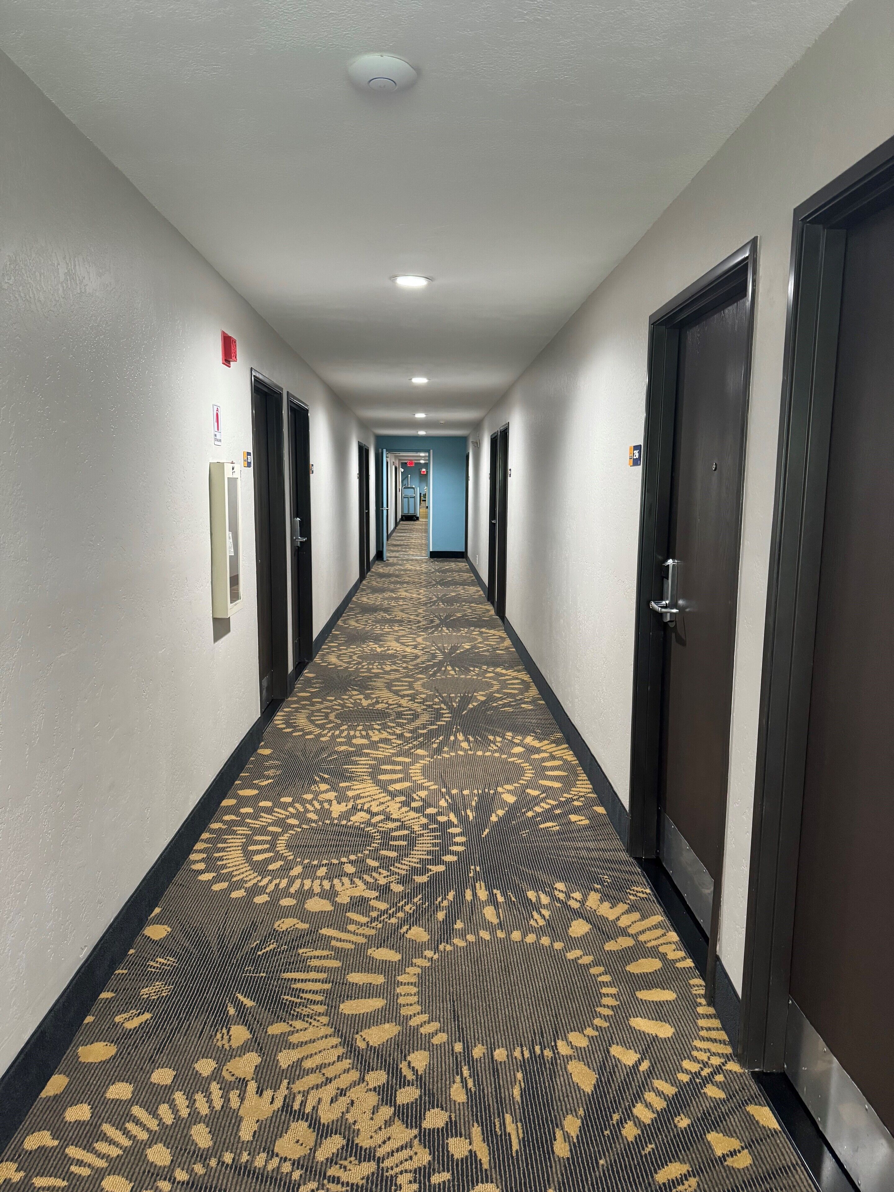 A hotel hallway with carpeted floors, doors on both sides, and a fire alarm on the left wall.
