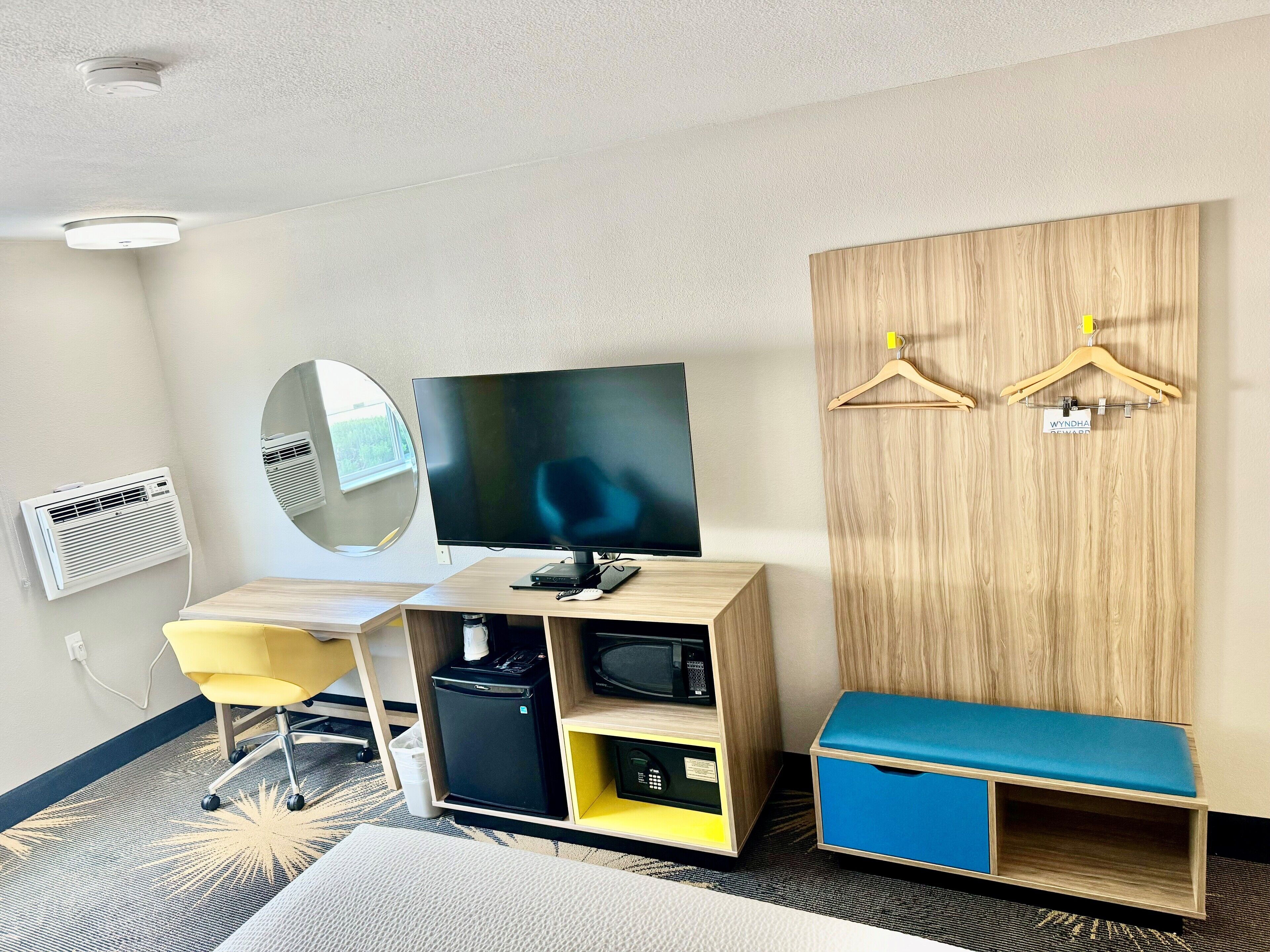 Habitació d'hotel amb televisió, mirall, nevera petita, un banc i armari amb penjalls.