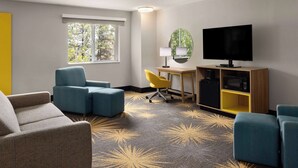 A hotel room with a large flat-screen TV, two blue armchairs, a yellow chair, a desk with a lamp, and a small cabinet with a microwave.