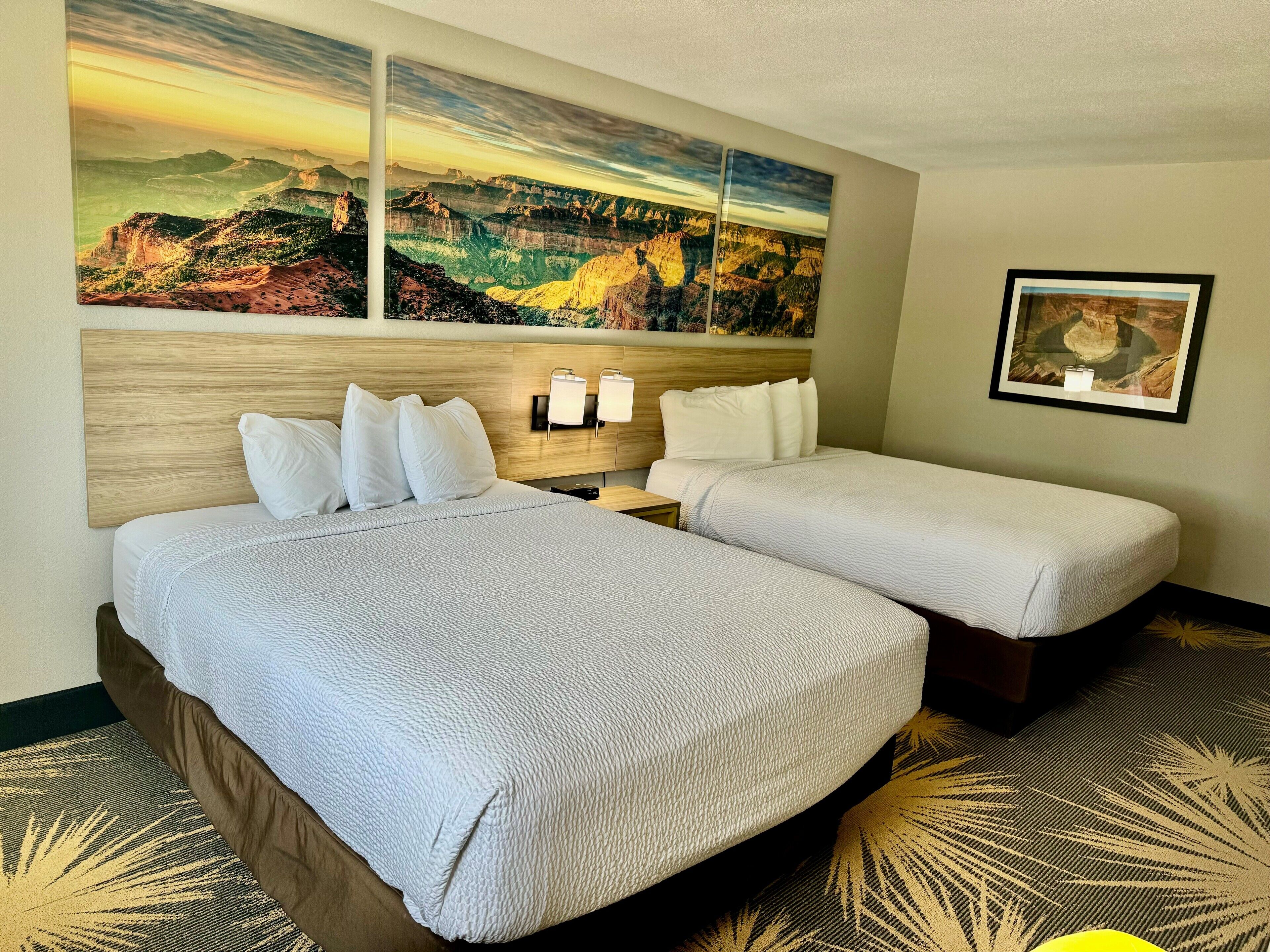 A hotel room with two beds, a wooden headboard, a nightstand with lamps, and a large landscape painting above the beds.