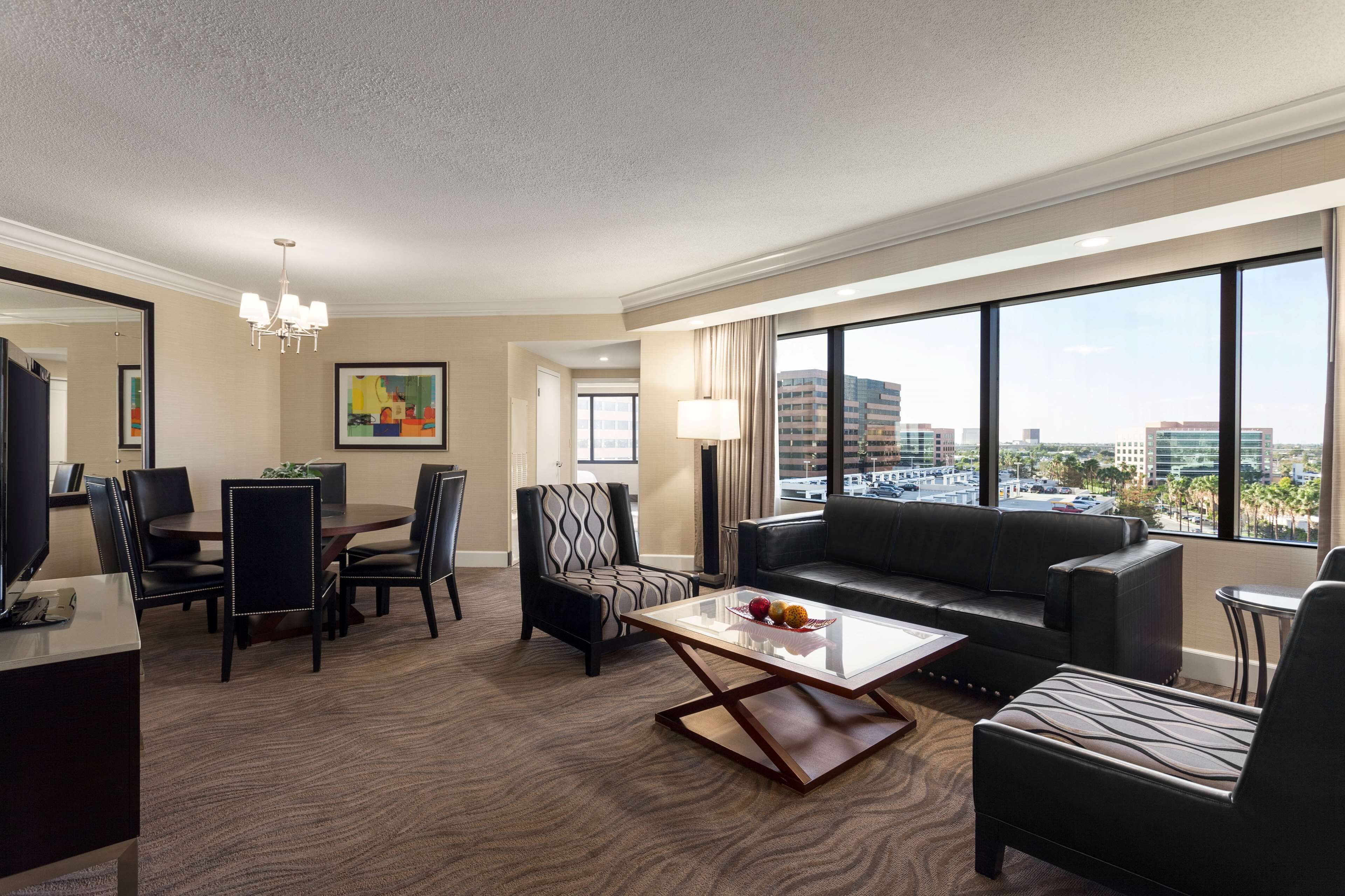 A hotel room with a dining area, a sofa, a coffee table, and large windows offering a city view.