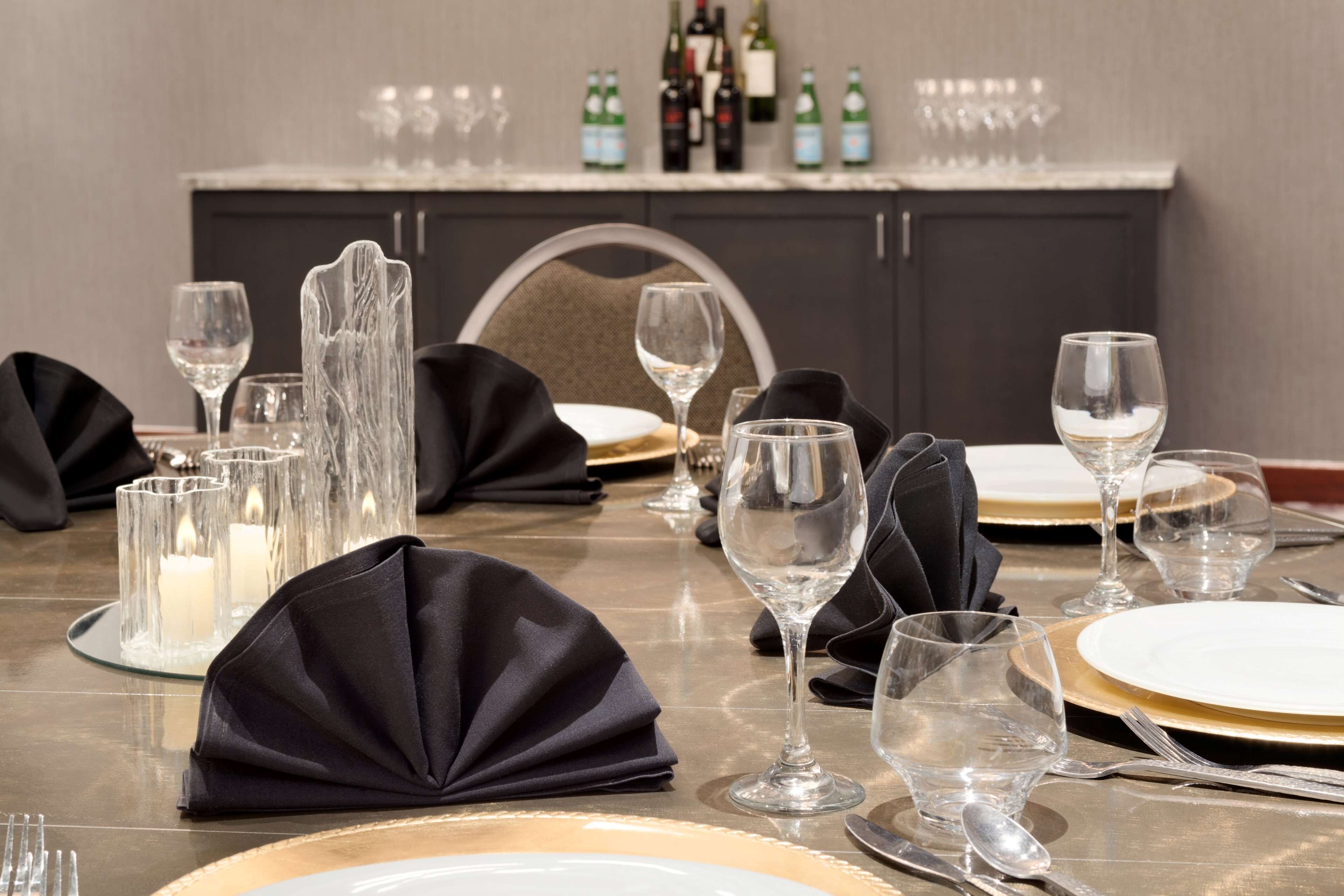 A dining table set with plates, glasses, and cutlery, with candles and bottles in the background.