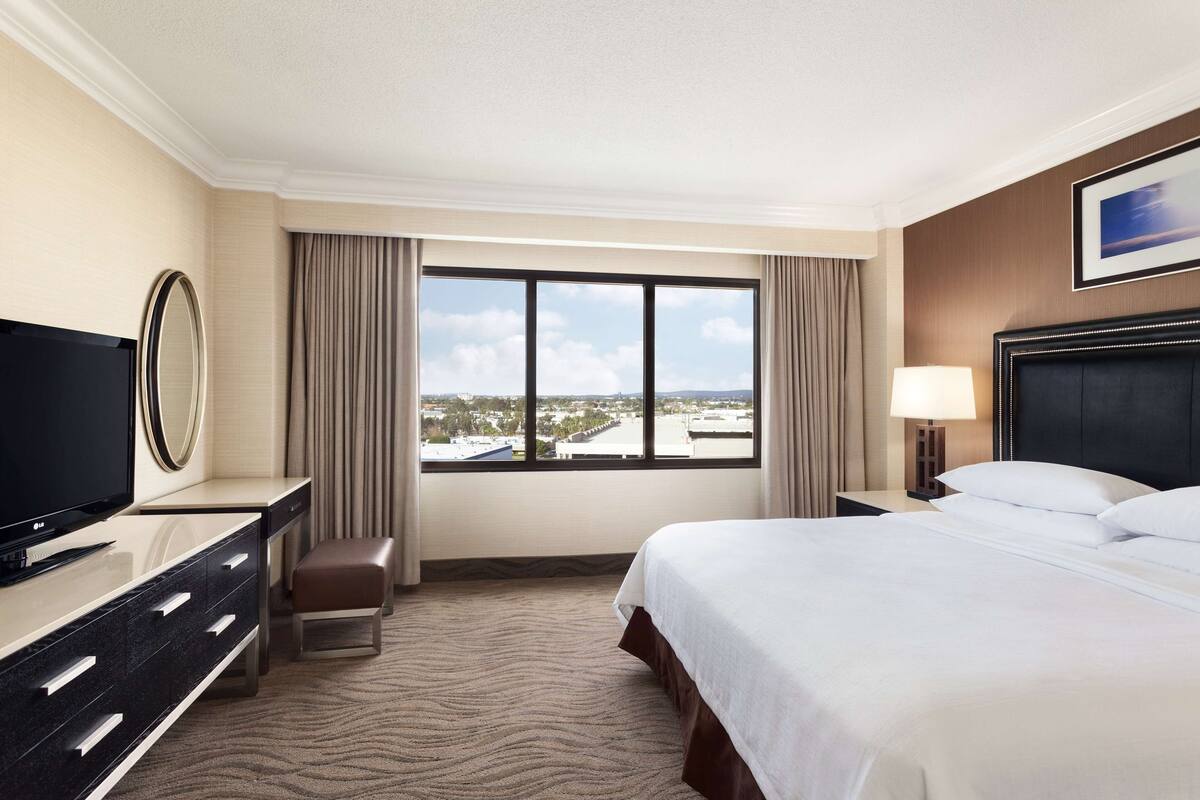 Ein Hotelzimmer mit Bett, Fernseher, Schreibtisch, Stuhl und einem großen Fenster mit Blick auf die Stadt.