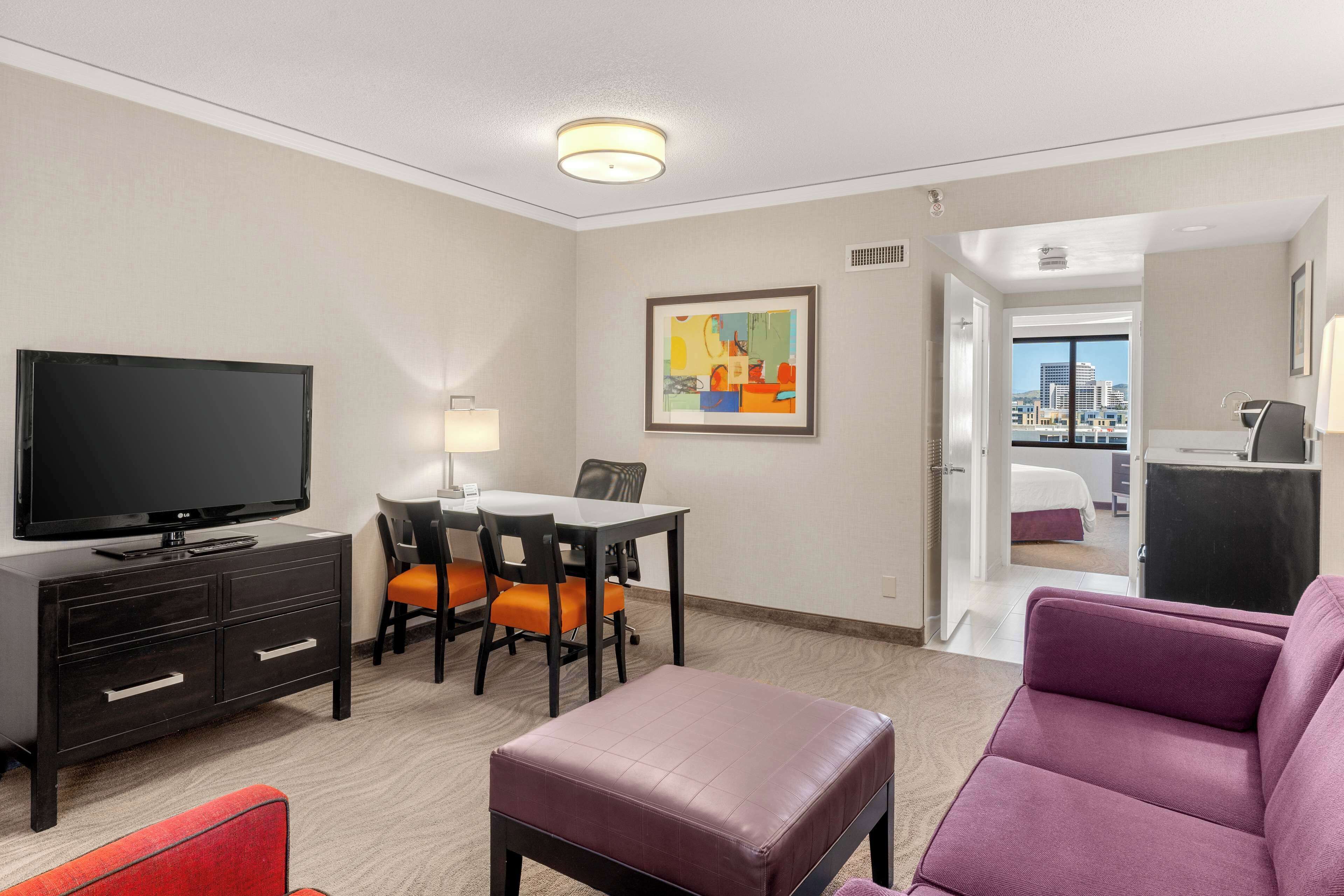 A hotel room with a TV, a dining table with chairs, a purple sofa, a red chair, and a city view through the door.