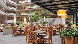 A spacious indoor area with multiple dining tables and chairs, surrounded by lush green plants and a modern architectural design with a mezzanine level. - Embassy Suites by Hilton Irvine Orange County Airport (Irvine)