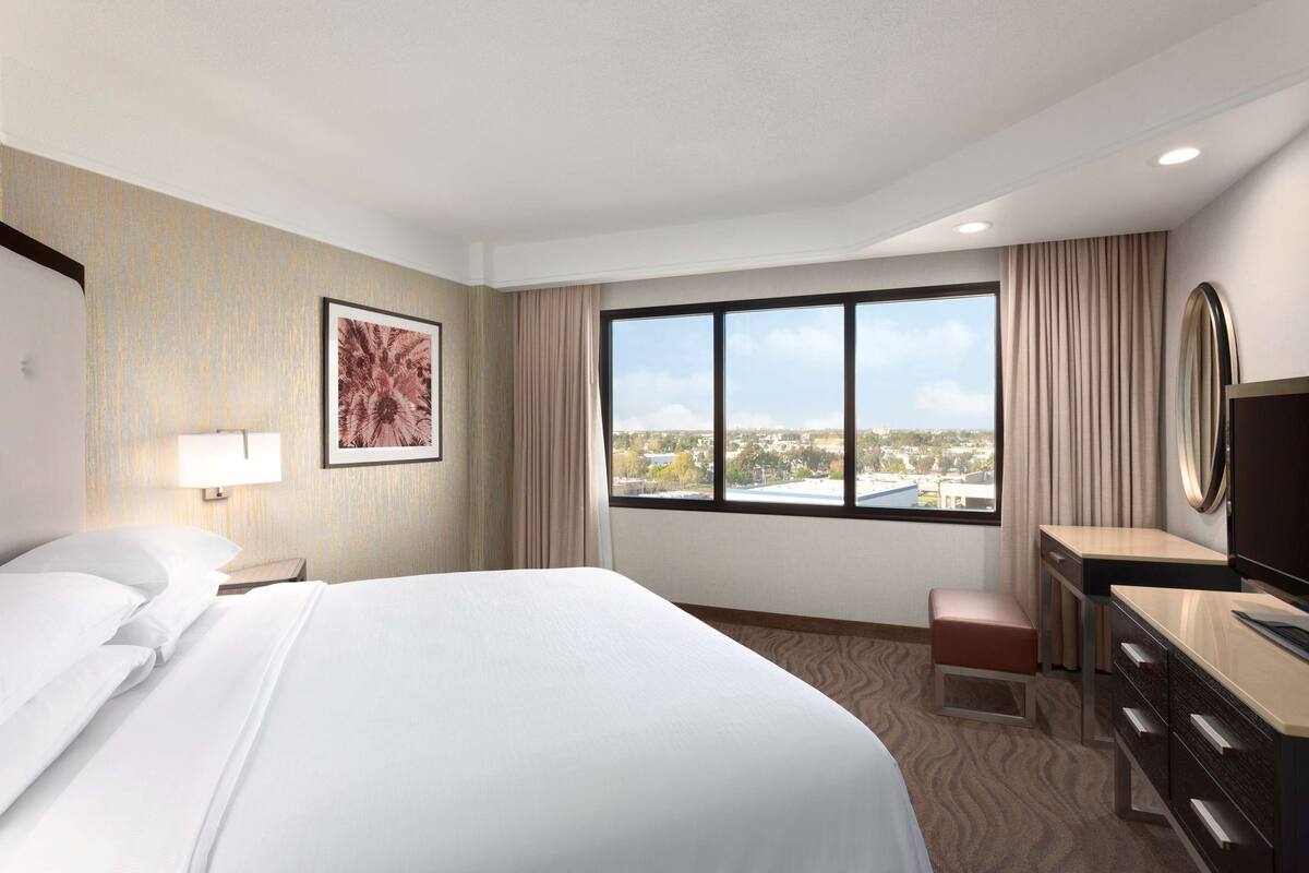 Ein Hotelzimmer mit einem großen Bett, einem Schreibtisch, einem Stuhl, einem Fernseher und einem Fenster mit Blick auf die Stadt.