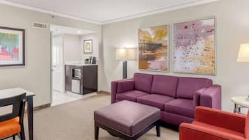 A modern hotel room with a purple sofa, a red armchair, a dining table with chairs, a kitchen area, and two large abstract paintings on the walls.