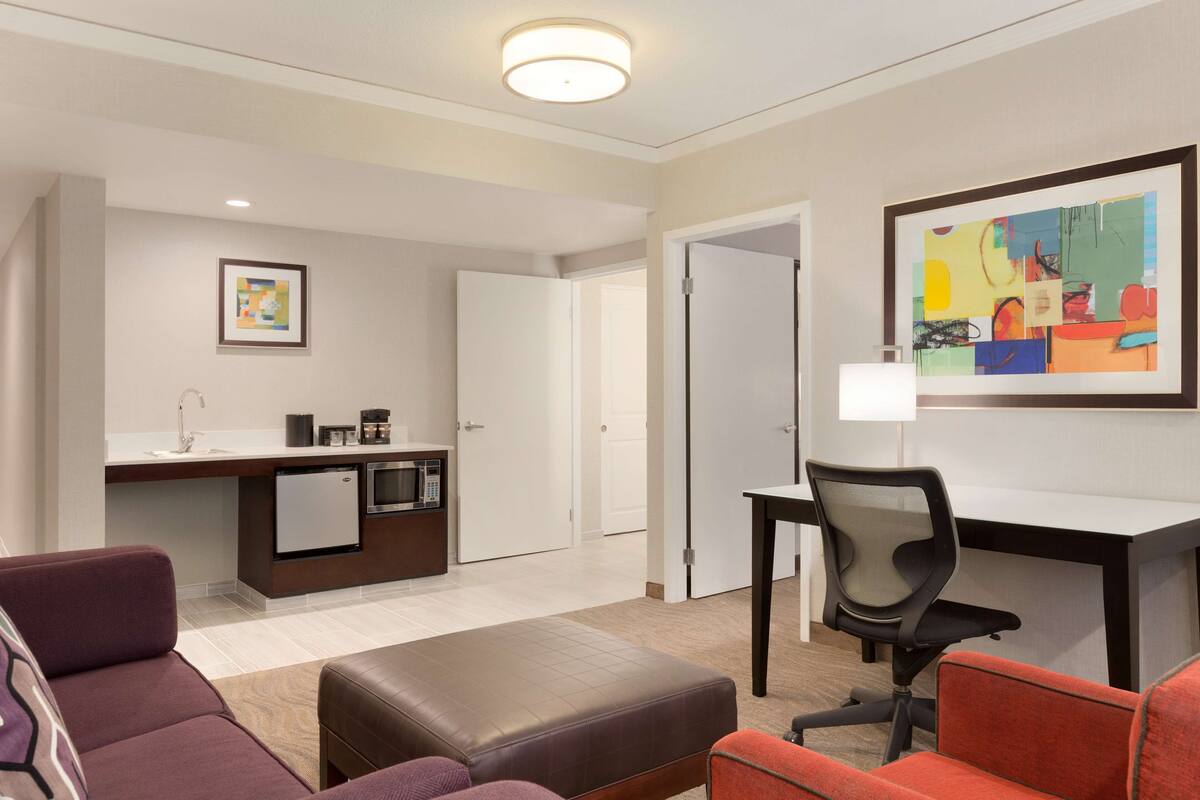 Ein modernes Hotelzimmer mit Küchenzeile, Essecke und Wohnbereich mit Sofa und Hocker.