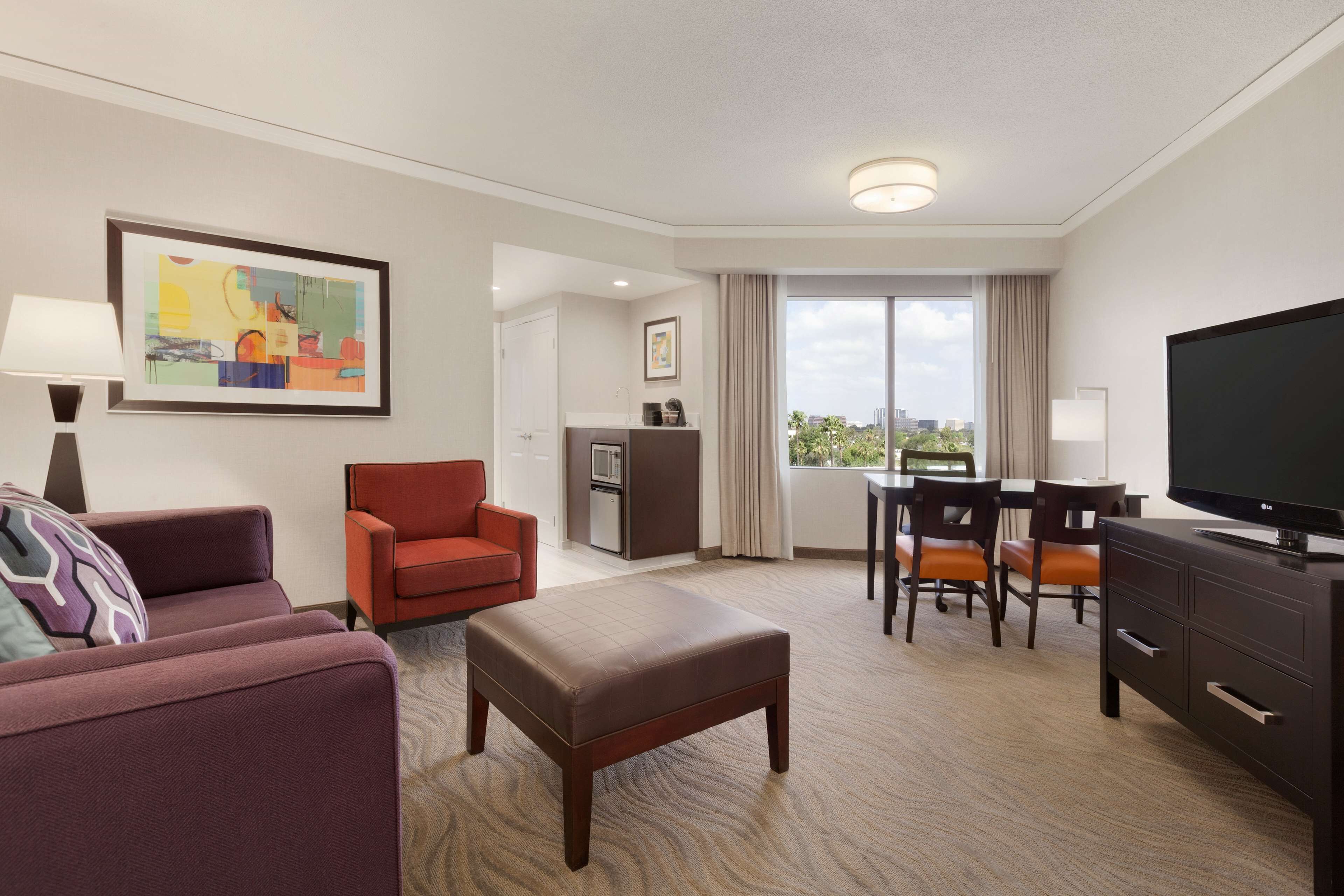Quarto de hotel com sofá, poltrona, banquinho, mesa de jantar com cadeiras, televisão e obras de arte na parede.