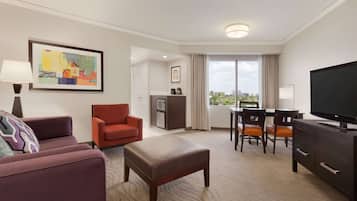 Quarto de hotel com sofá, poltrona, banquinho, mesa de jantar com cadeiras, televisão e obras de arte na parede.