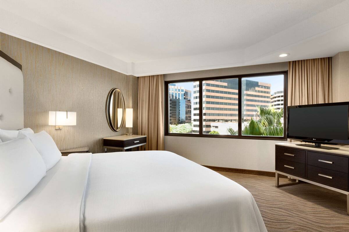 Ein Hotelzimmer mit einem großen Bett, einem Fernseher, einem Schreibtisch und einem Fenster mit Blick auf die Stadt.
