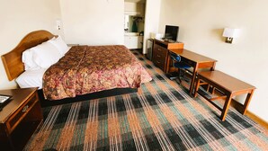 Quarto de hotel com cama, escrivaninha, cadeira, televisão e luminária.