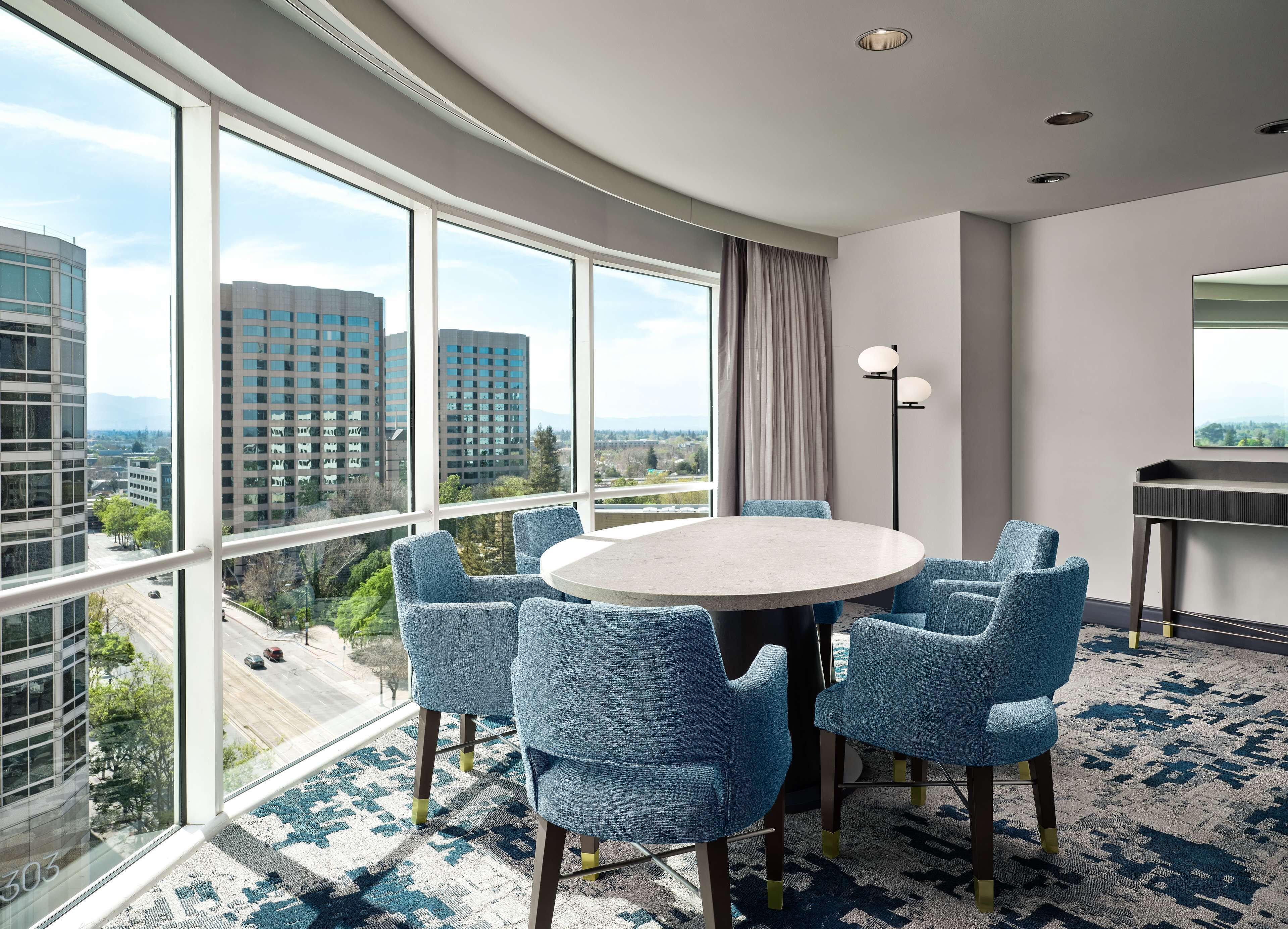 Moderni hotellihuone, jossa on suuri ikkuna, pyöreä pöytä tuoleineen, lattiavalaisin ja pieni työpöytä.