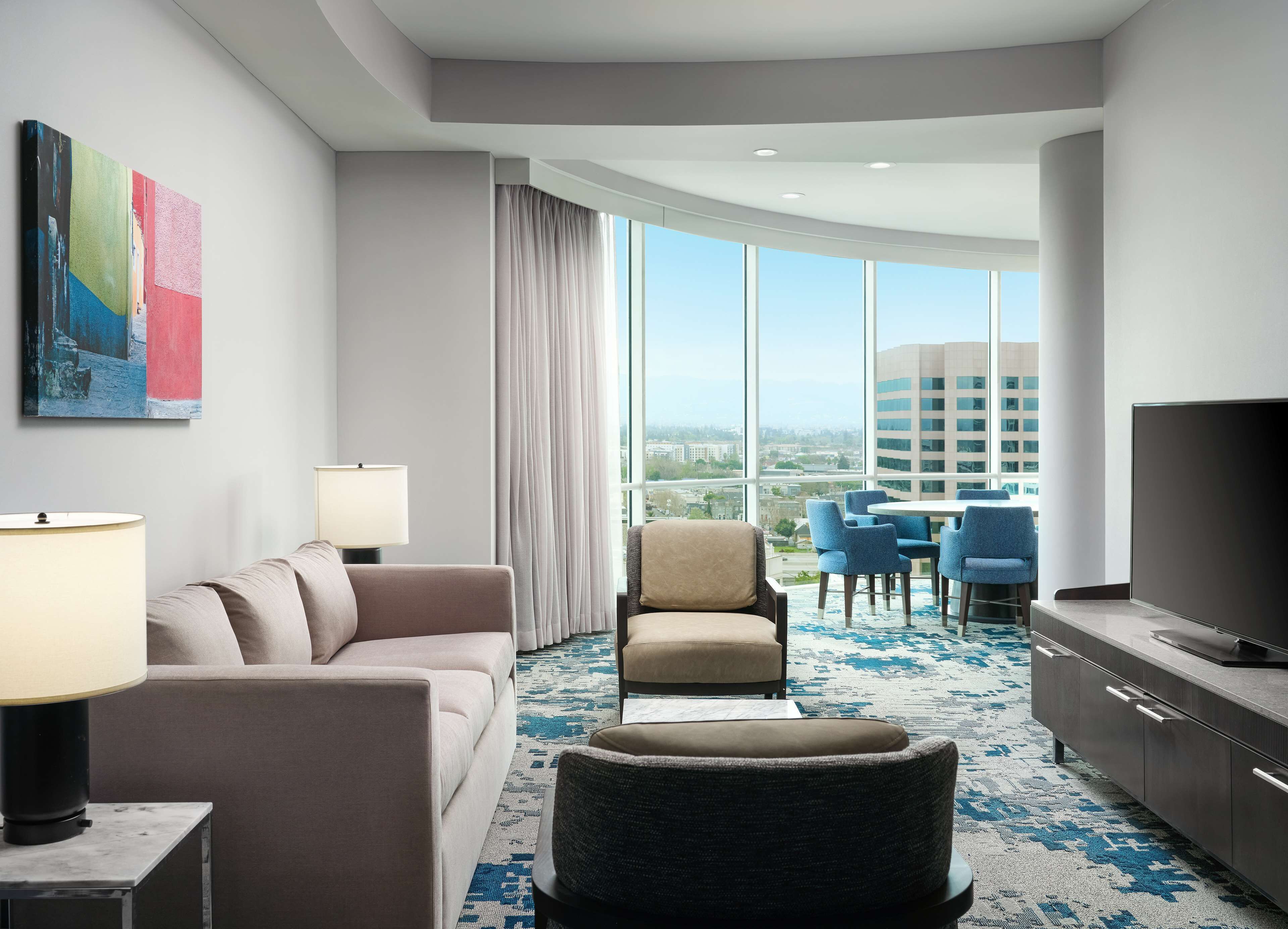 Une chambre d’hôtel moderne dotée d’une grande fenêtre, d’un canapé, d’un fauteuil, d’une télévision et d’un coin repas avec des chaises et une table.