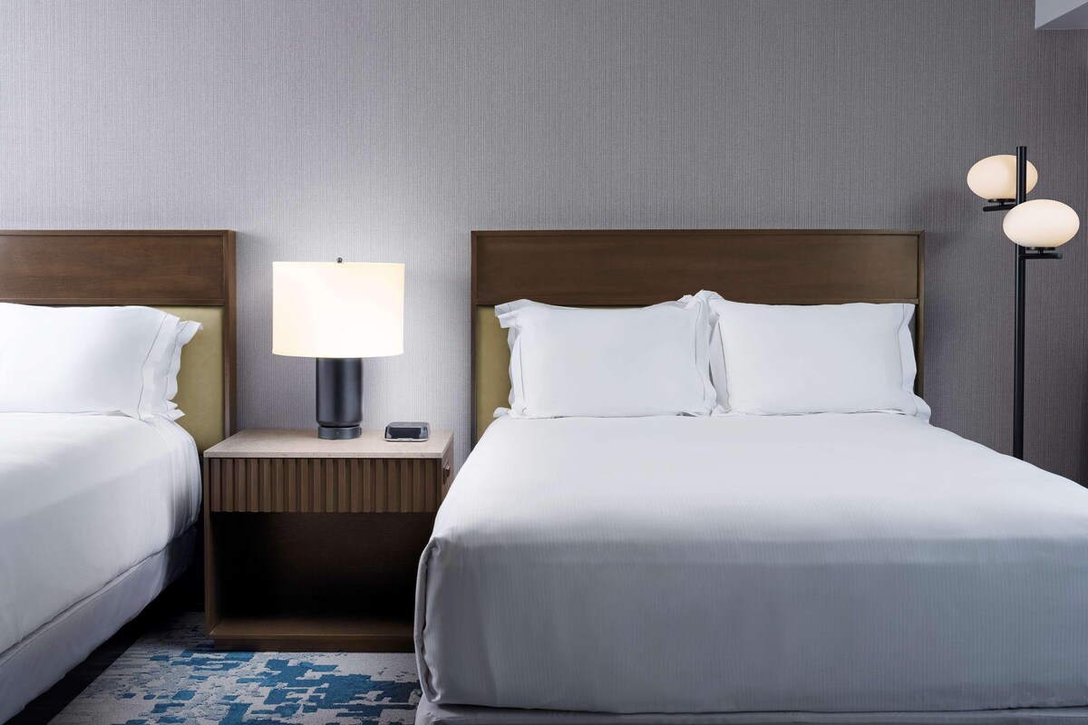 A hotel room with two beds, a nightstand with a lamp, a wall-mounted light, and a patterned rug.