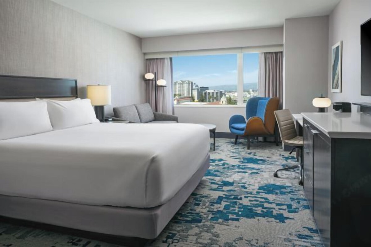 A hotel room with a large bed, a desk, a chair, and a view of the city through the window.
