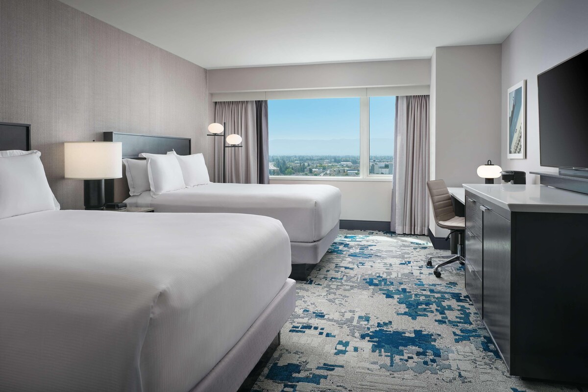 A hotel room with two beds, a desk, a chair, a TV, and a large window with a view of the city.