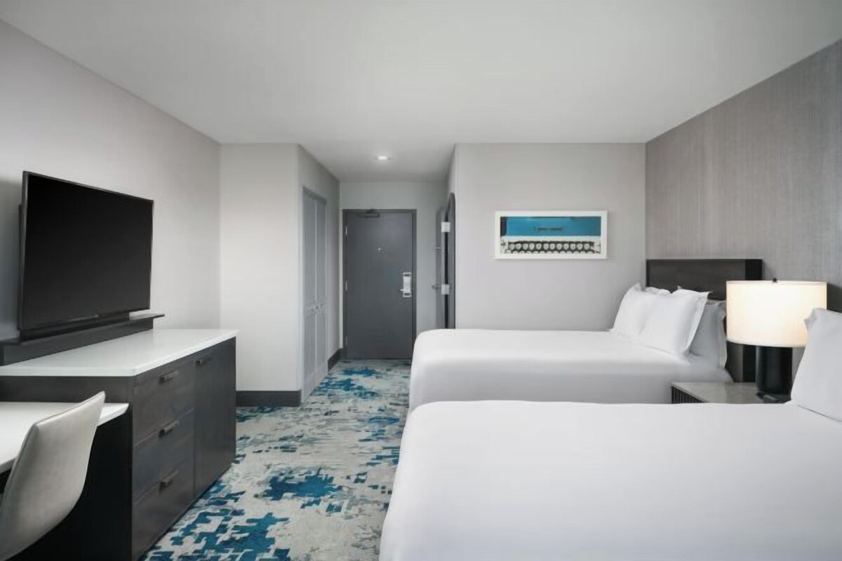 A hotel room with a bed, a desk with a TV, a chair, and a nightstand with a lamp.