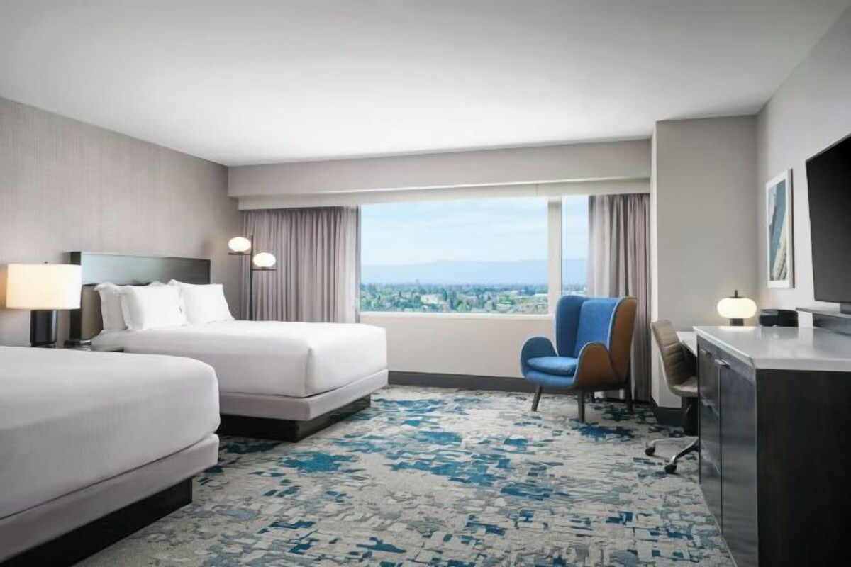 A hotel room with two beds, a desk, a chair, and a large window offering a city view.