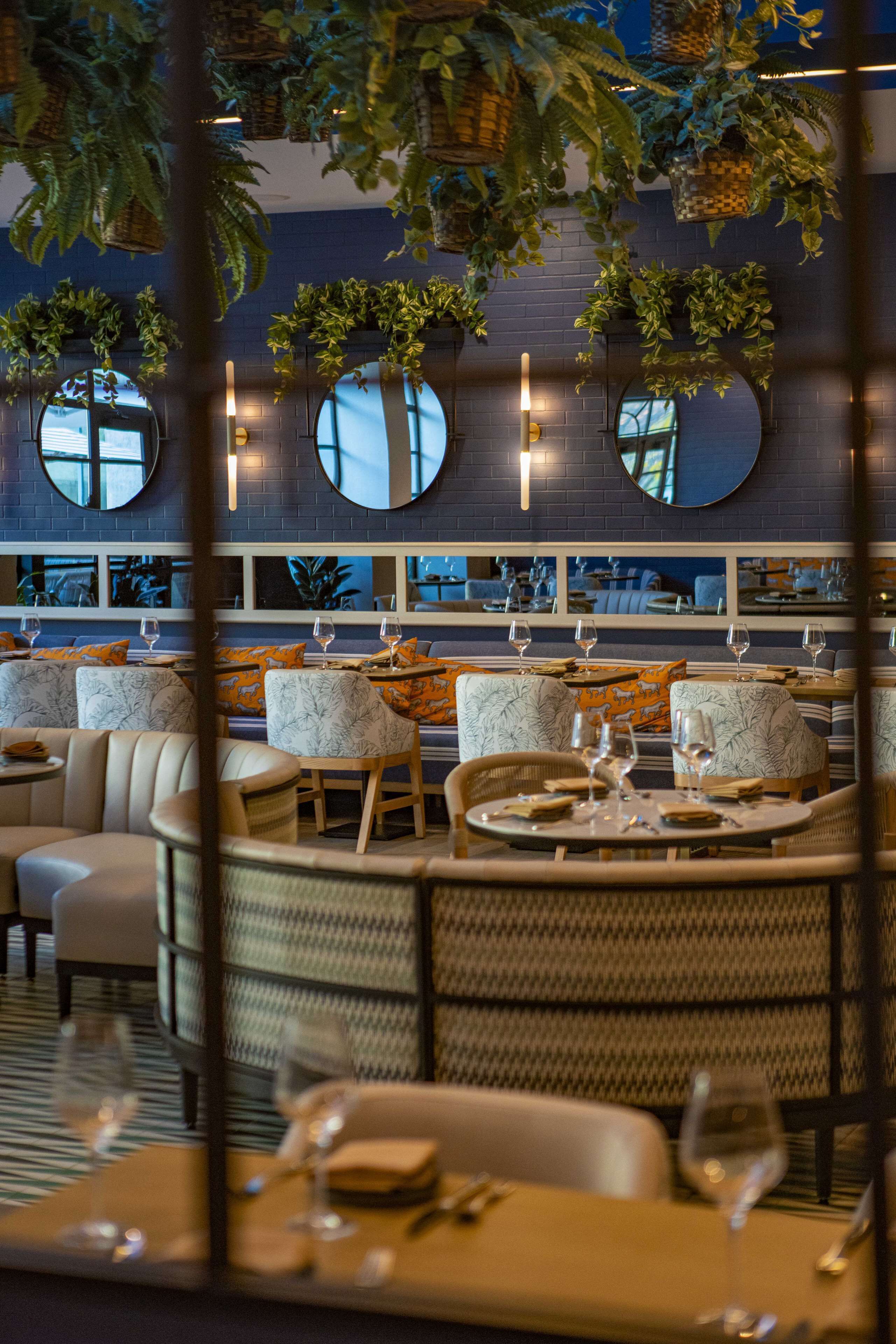 A restaurant with round tables and chairs, some with cushions. There are hanging plants and a brick wall with mirrors.