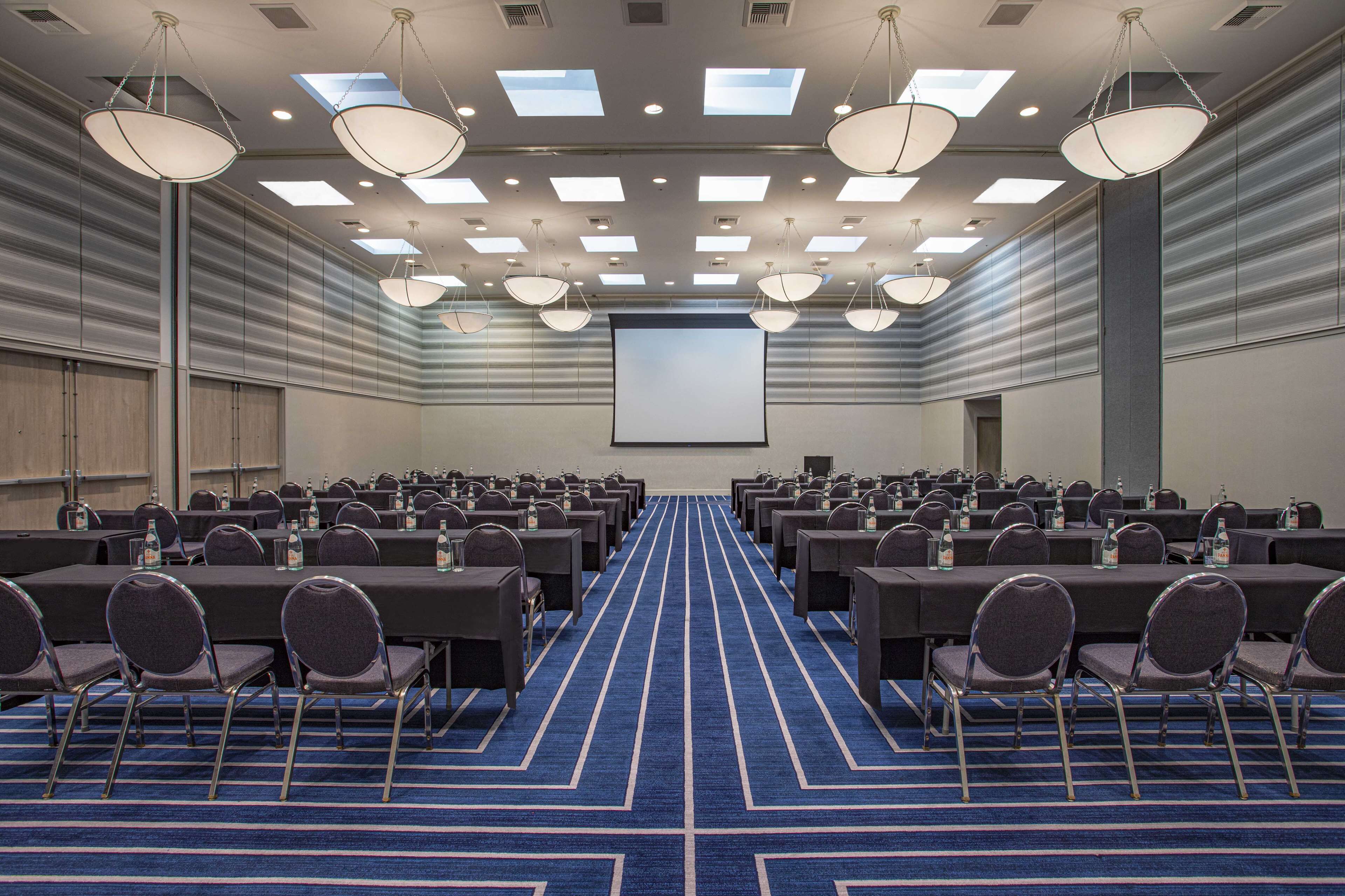 Uma sala de conferências com mesas e cadeiras dispostas em fileiras, uma tela grande e um tapete azul.