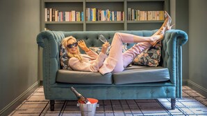 A teal sofa with a floral pillow, a champagne bottle, and a book-filled bookshelf in the background.