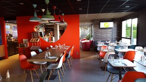 A restaurant with a modern interior, featuring a mix of red and neutral tones, multiple dining tables, and colorful chairs. - Hôtel ibis Cambrai (Fontaine-Notre-Dame)