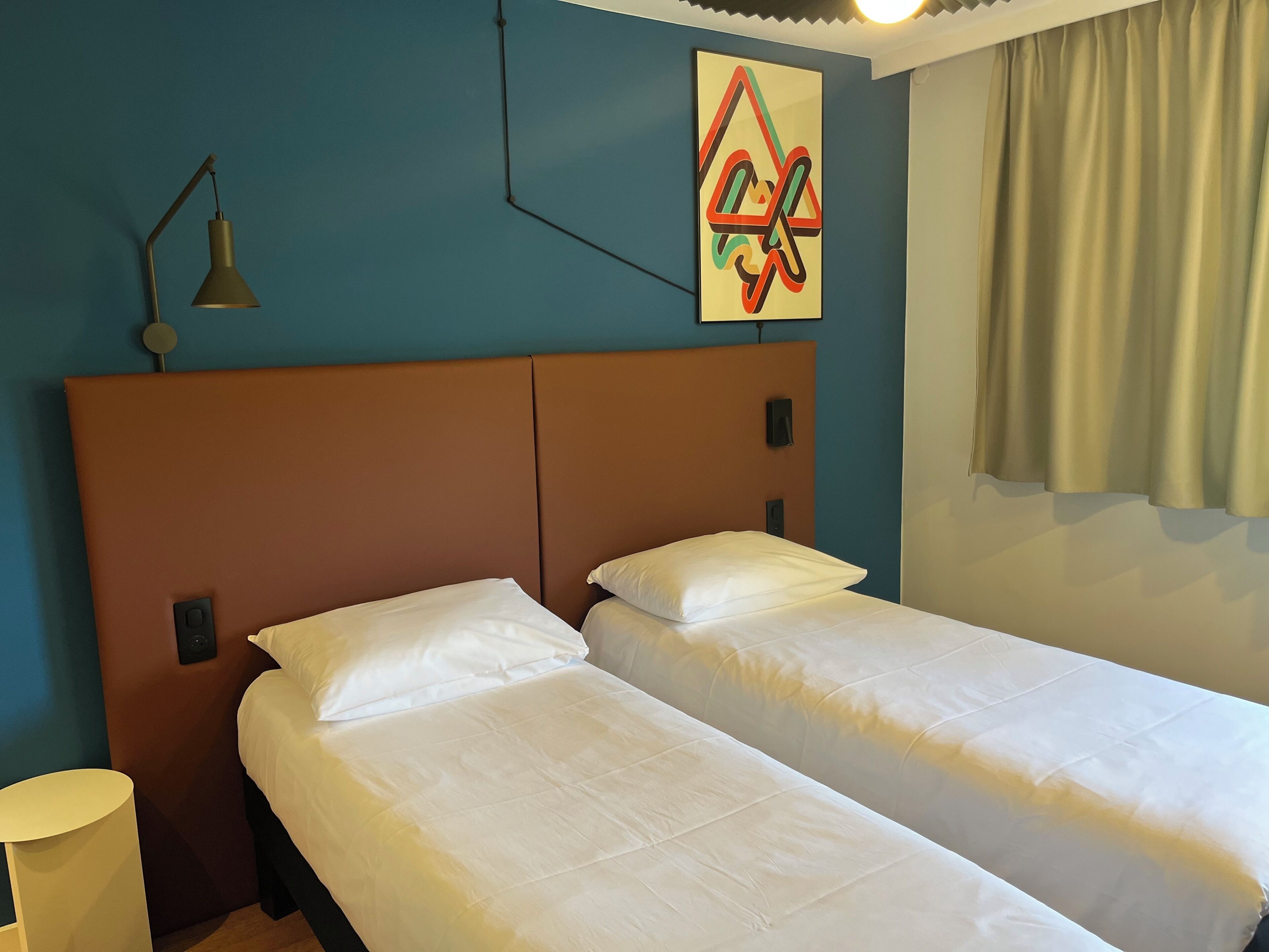 Une chambre avec deux lits simples, une table de chevet, une lampe fixée au mur et un tableau.