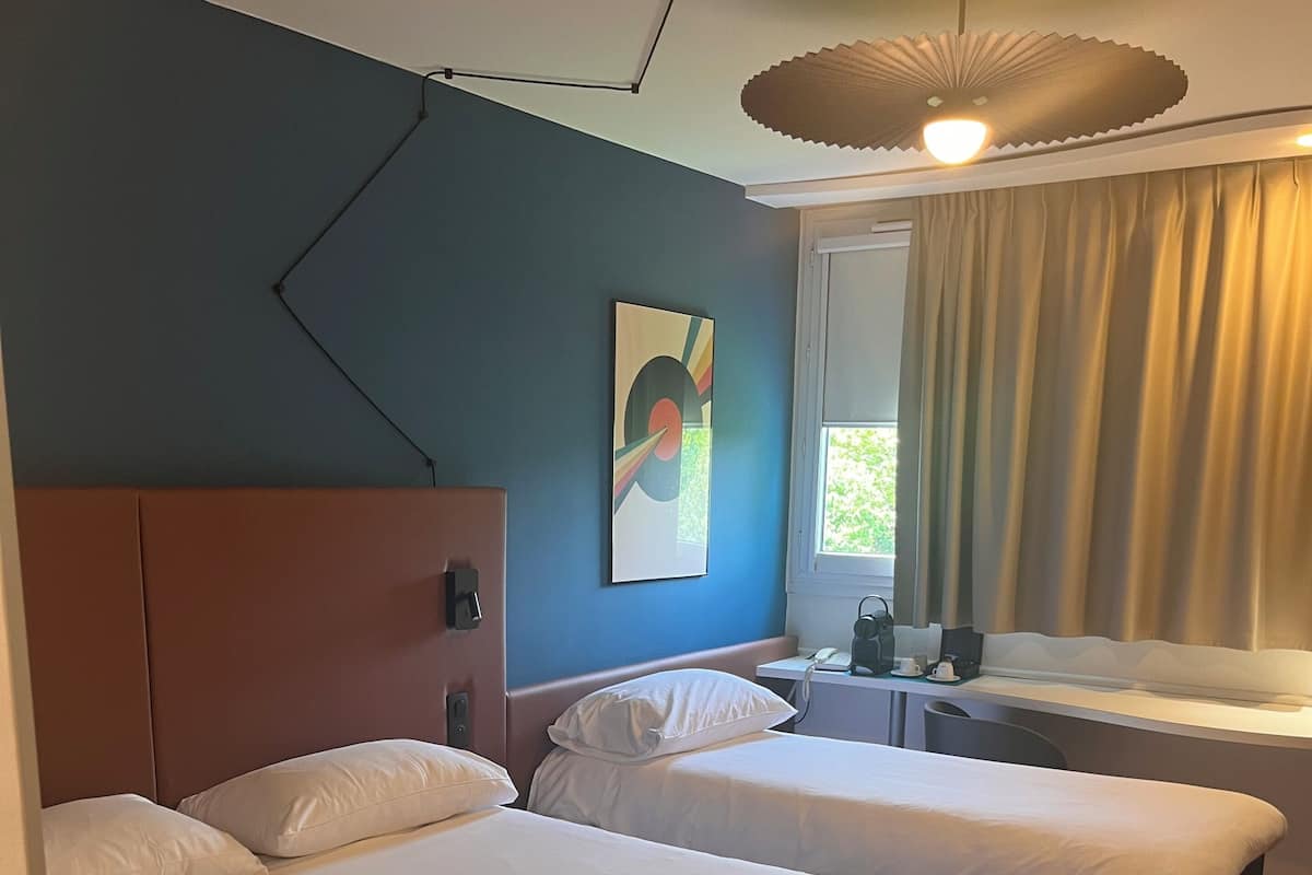 A hotel room with two beds, a window with curtains, a bedside table with a lamp, and a ceiling light fixture.