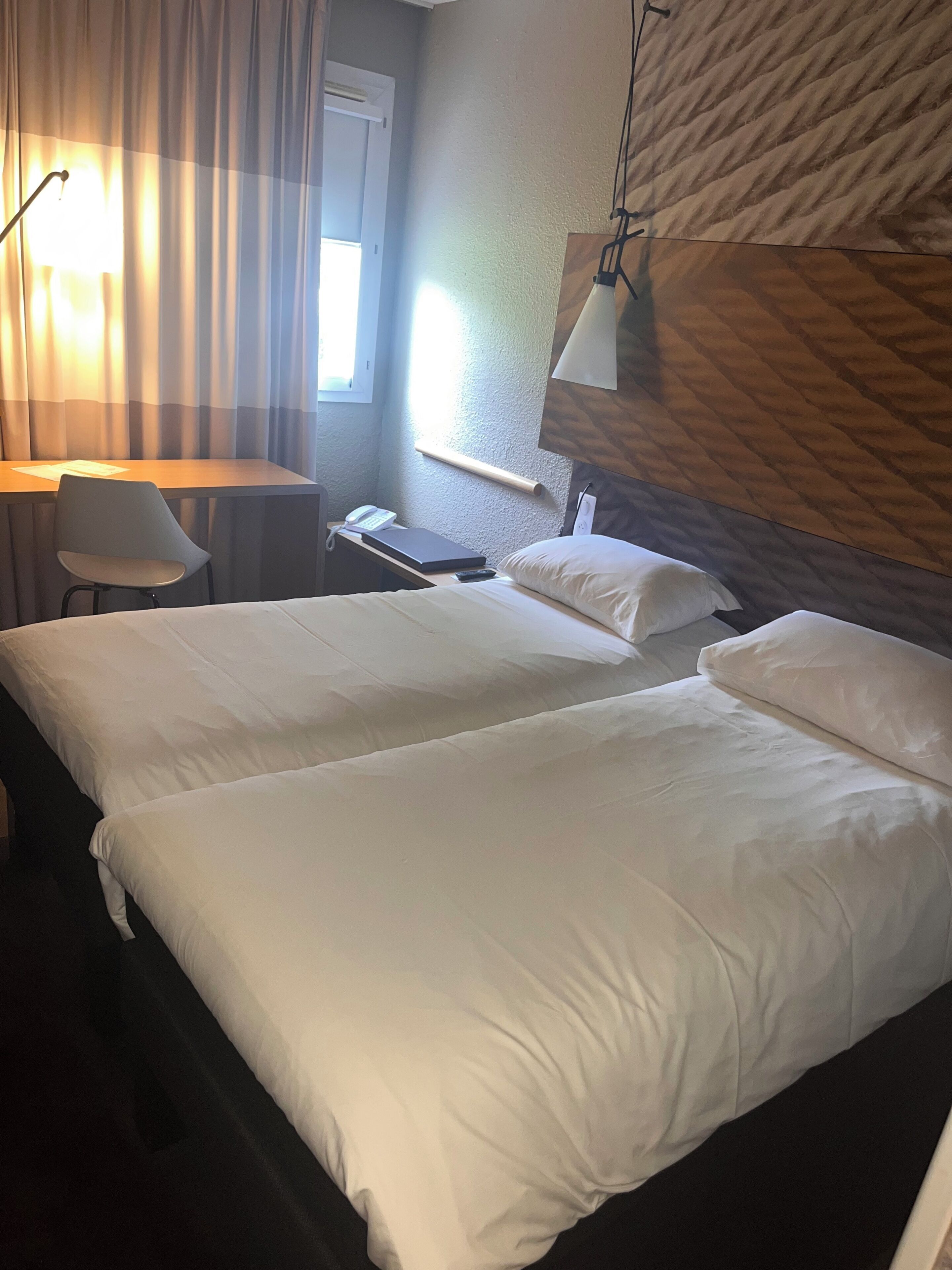 Ein Hotelzimmer mit zwei Betten, einem Schreibtisch, einem Stuhl, einer Lampe und einem fenster mit Vorhängen.