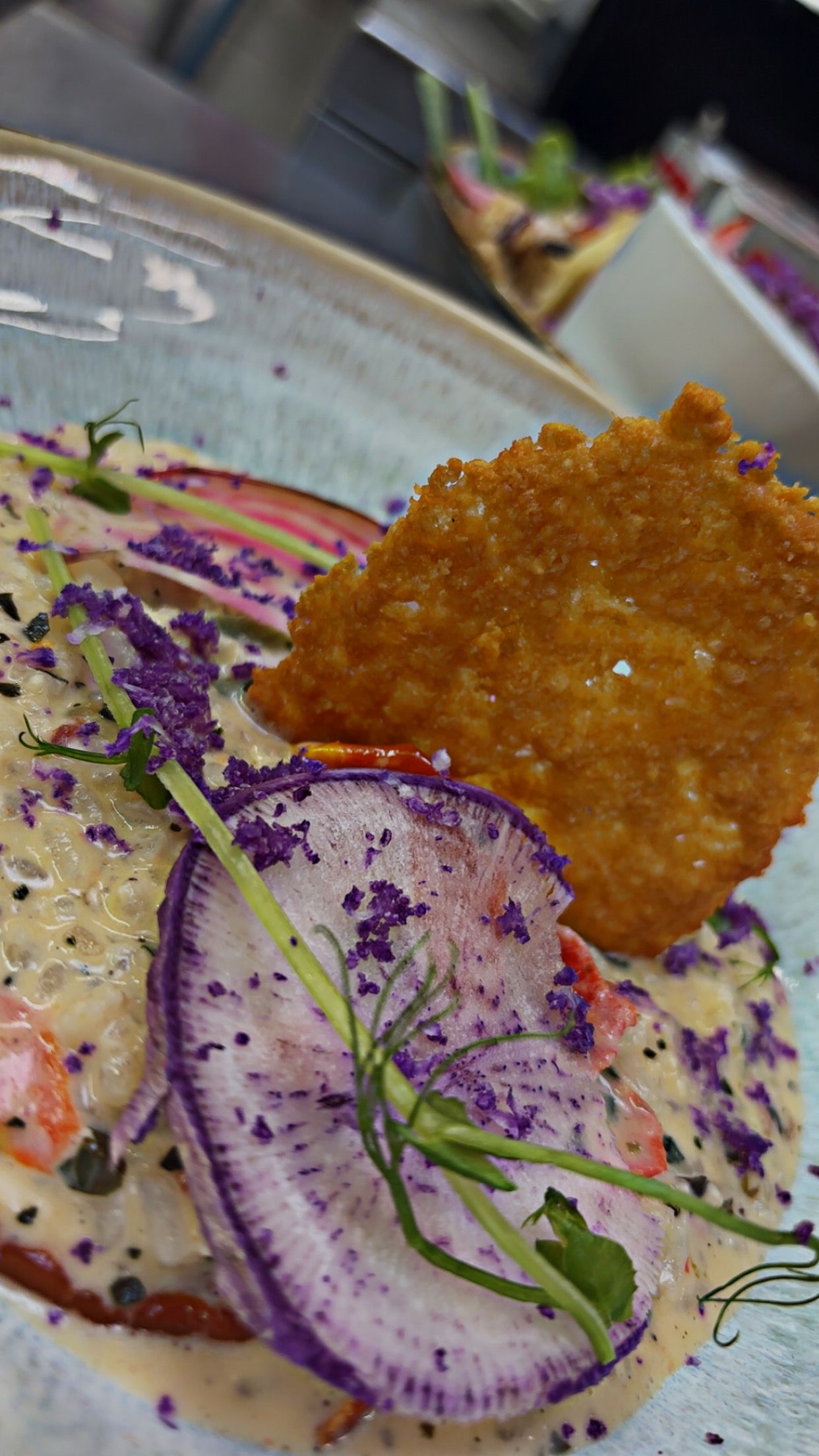 A close-up of a dish with a crispy item, possibly a fried item, and a creamy sauce with purple edible flowers and green garnish.