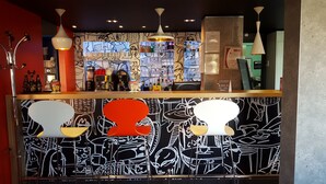 A bar area with a counter, bar stools, and a wall with abstract black and white patterns. - Hôtel ibis Cambrai (Fontaine-Notre-Dame)