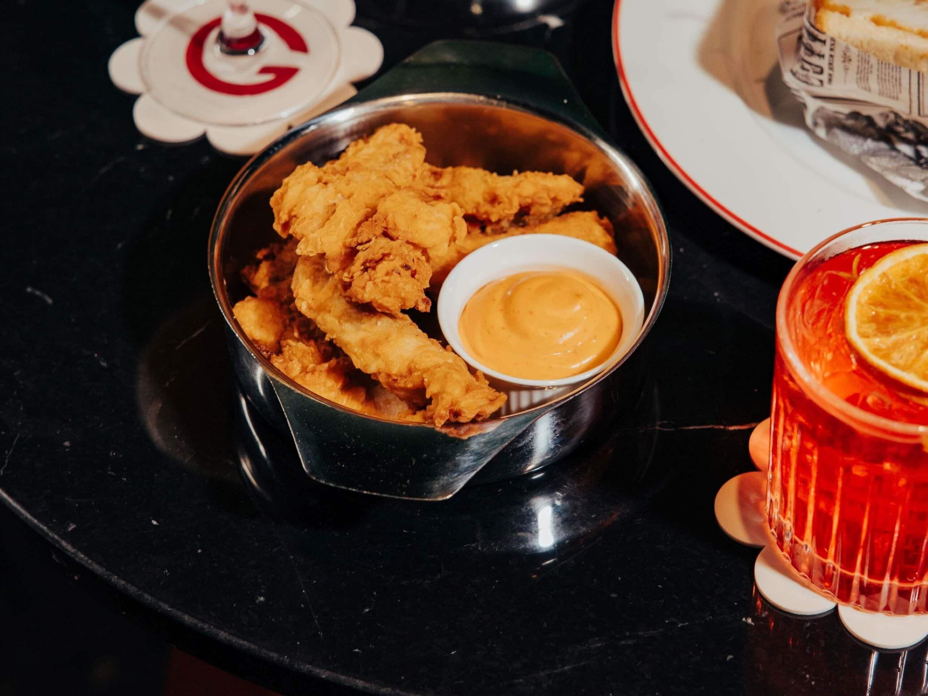 Un bol en métal rempli de poulet frit, accompagné d’une sauce, un verre d’une boisson rouge et une assiette avec un sandwich.