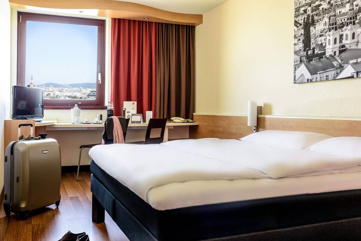 Quarto de hotel com cama, escrivaninha, cadeira, televisão e janela com cortinas.