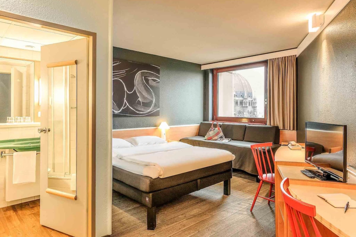 A hotel room with a bed, a desk, a chair, a window with a view of a building, and a large abstract painting on the wall.