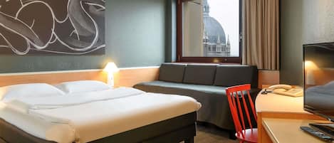 A hotel room with a bed, a sofa, a desk with a chair, a TV, and a window with a cityscape view.