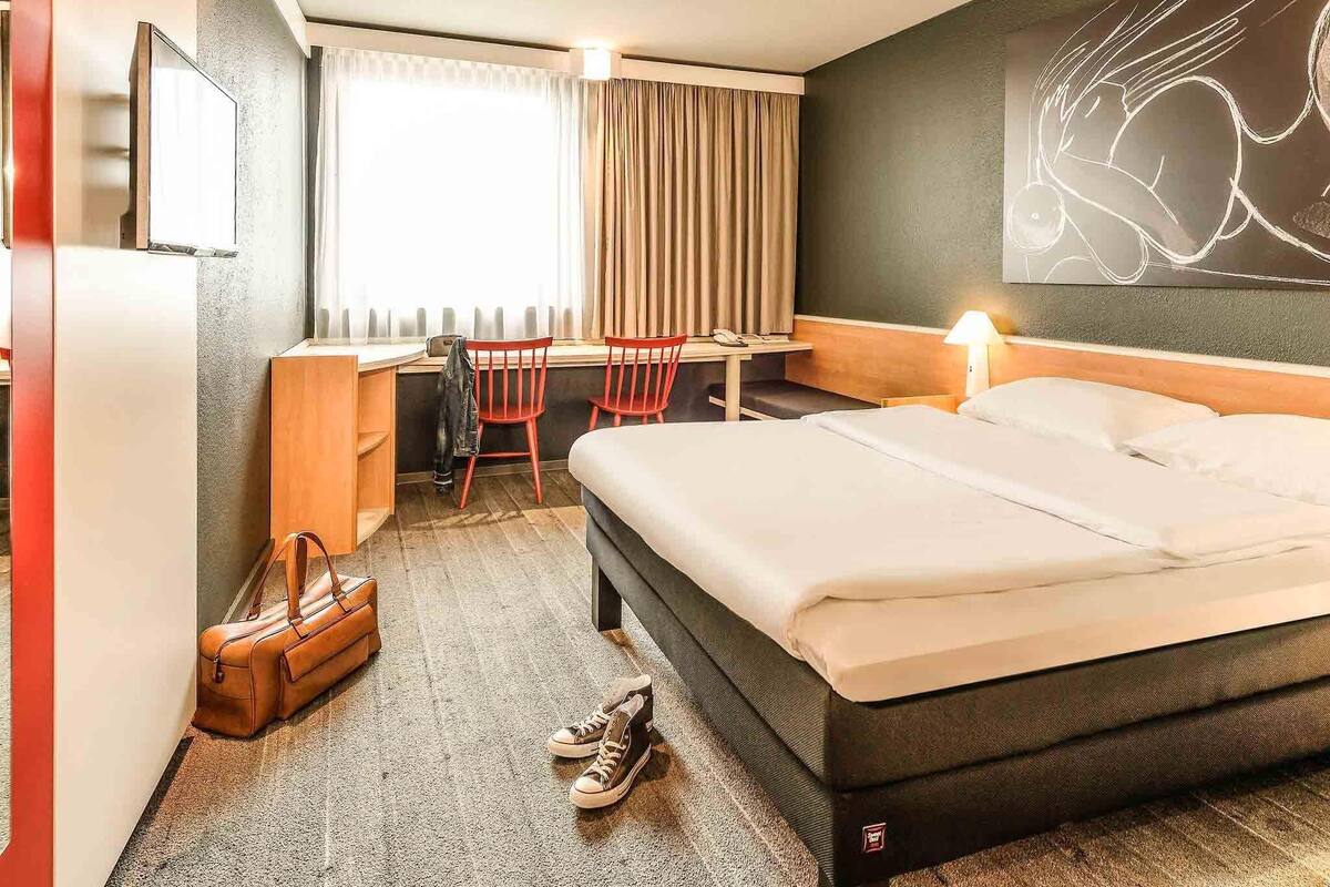 Quarto de hotel com cama, escrivaninha, cadeiras, abajur e um quadro na parede.