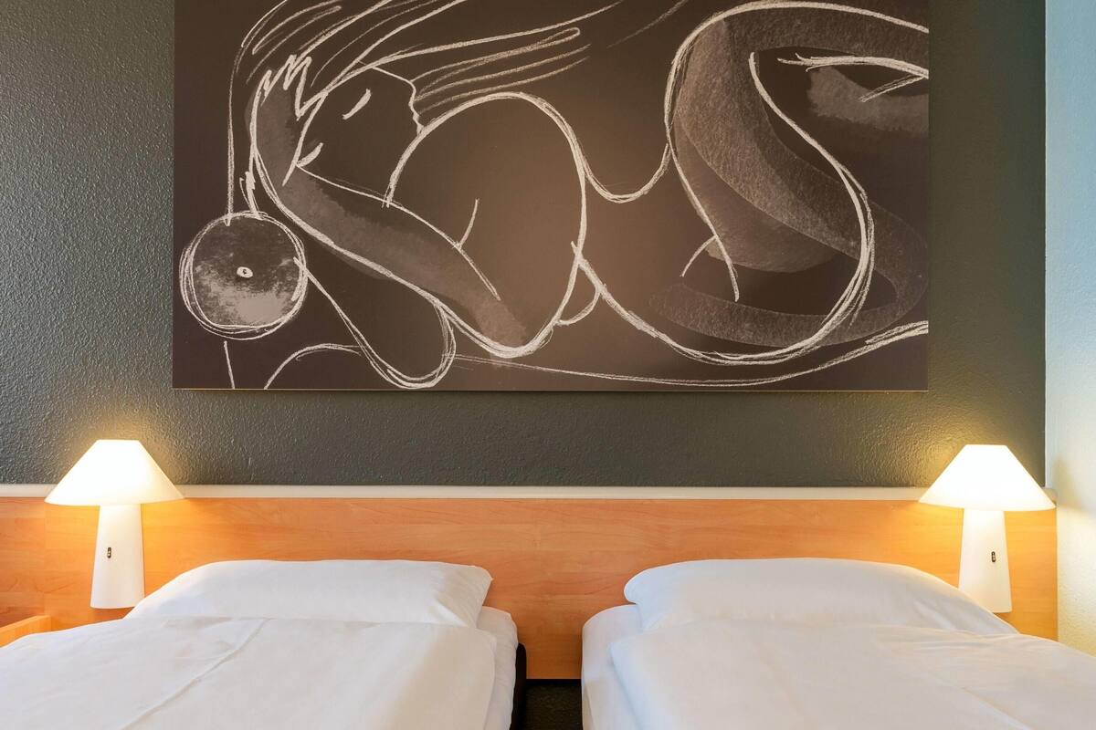 Quarto de hotel com duas camas, uma pintura abstrata grande na parede e duas luminárias de cabeceira.