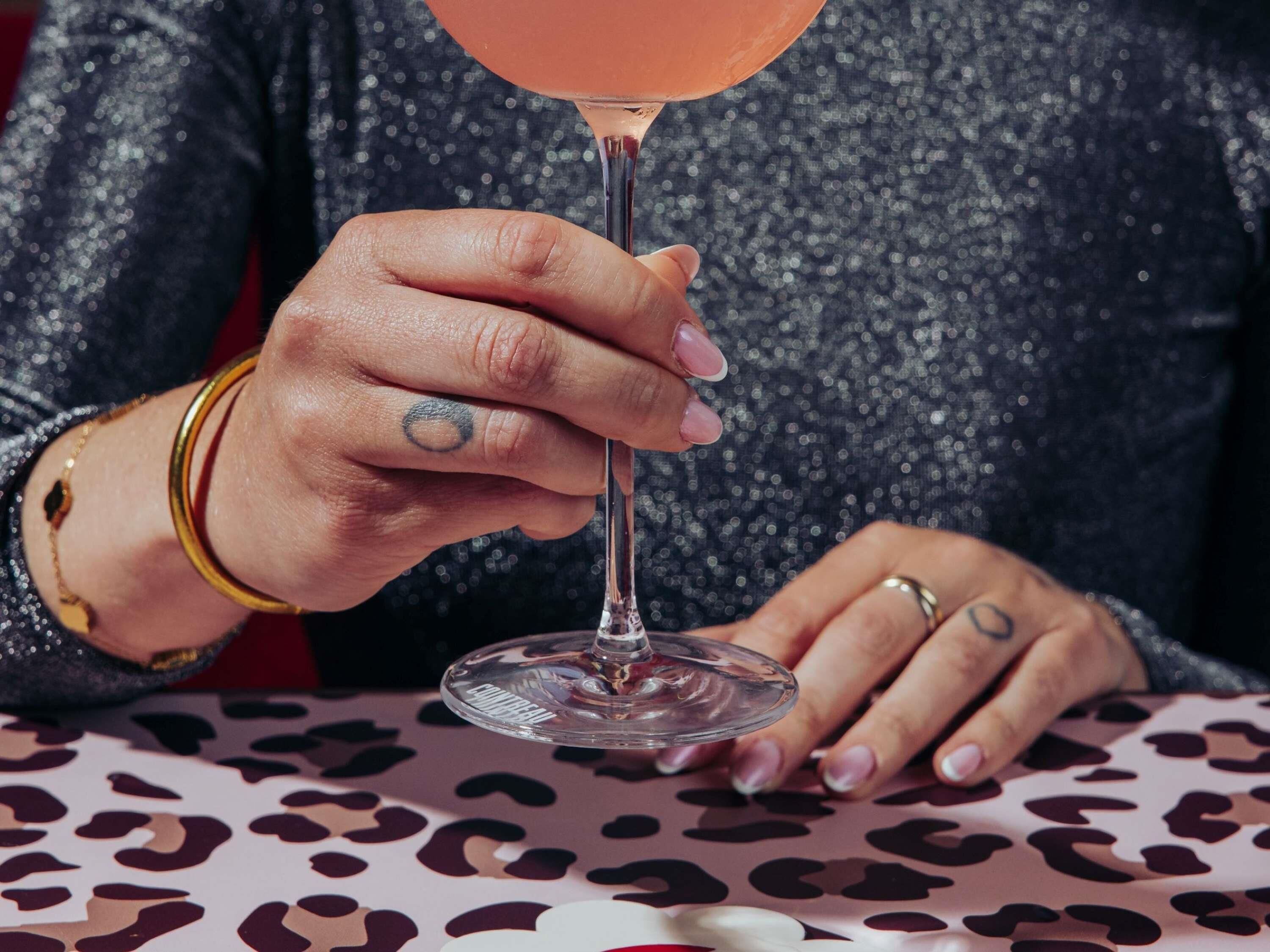 Una persona sosteniendo un vaso transparente con una bebida rosada, vistiendo una blusa brillante y una pulsera.