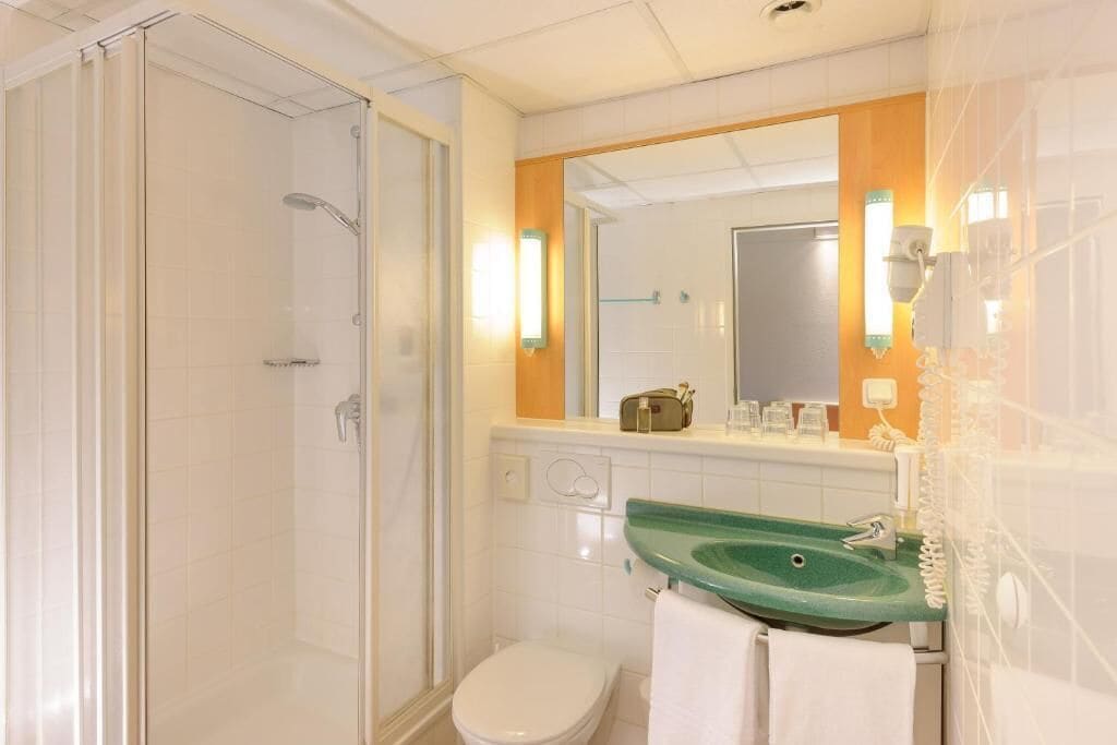 A bathroom with a glass shower enclosure, a white toilet, a green sink, and a mirror with a wooden frame.